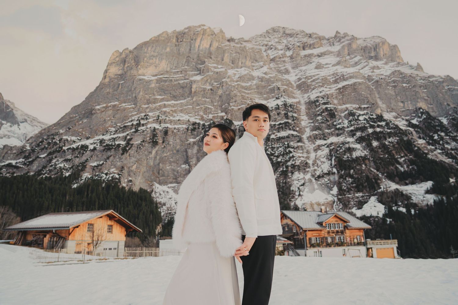 Elegant couple in white outfits standing back-to-back on snow with alpine lodge and mountain view.