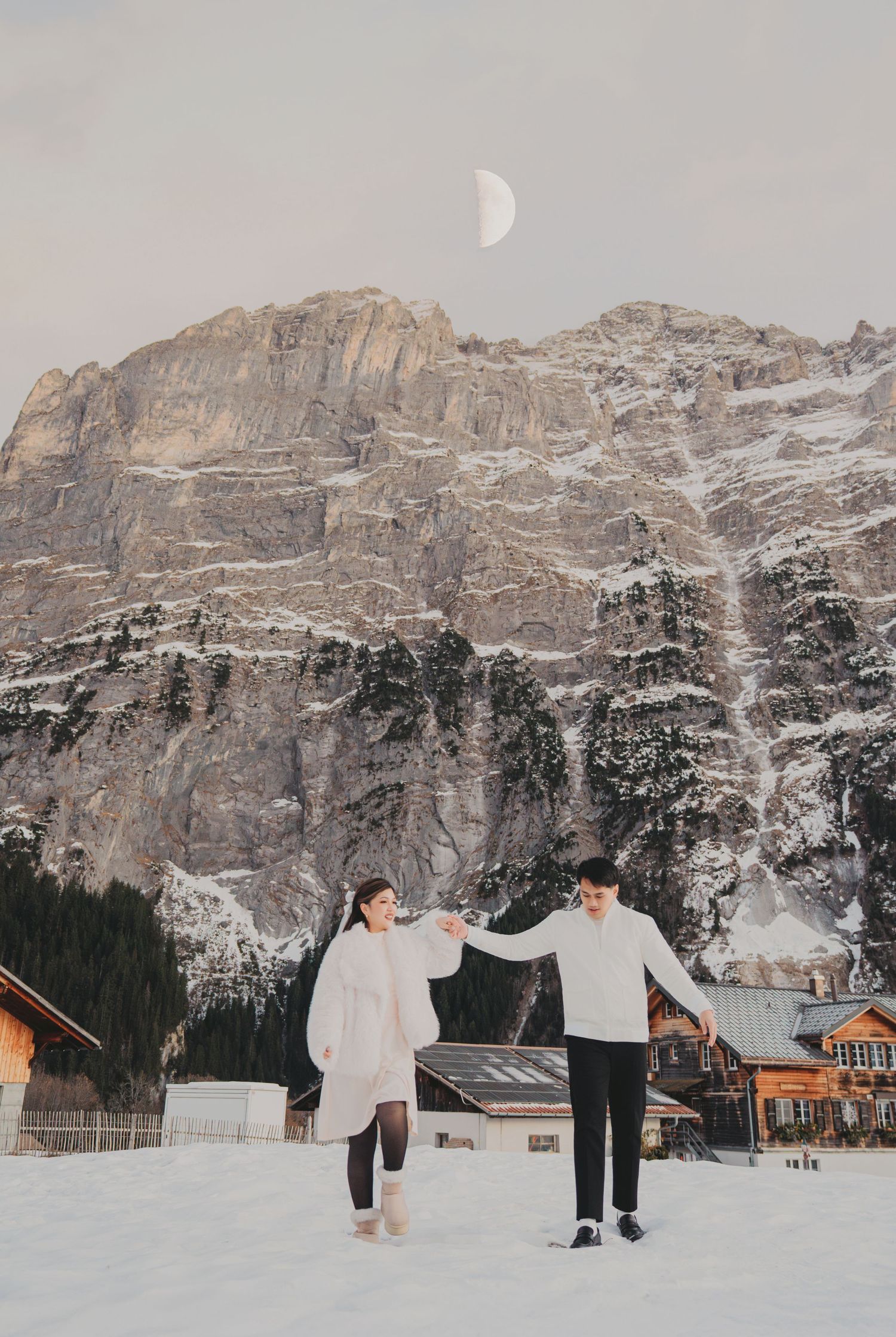 Couple holding hands in snowy Alpine landscape with half moon and rocky mountain.