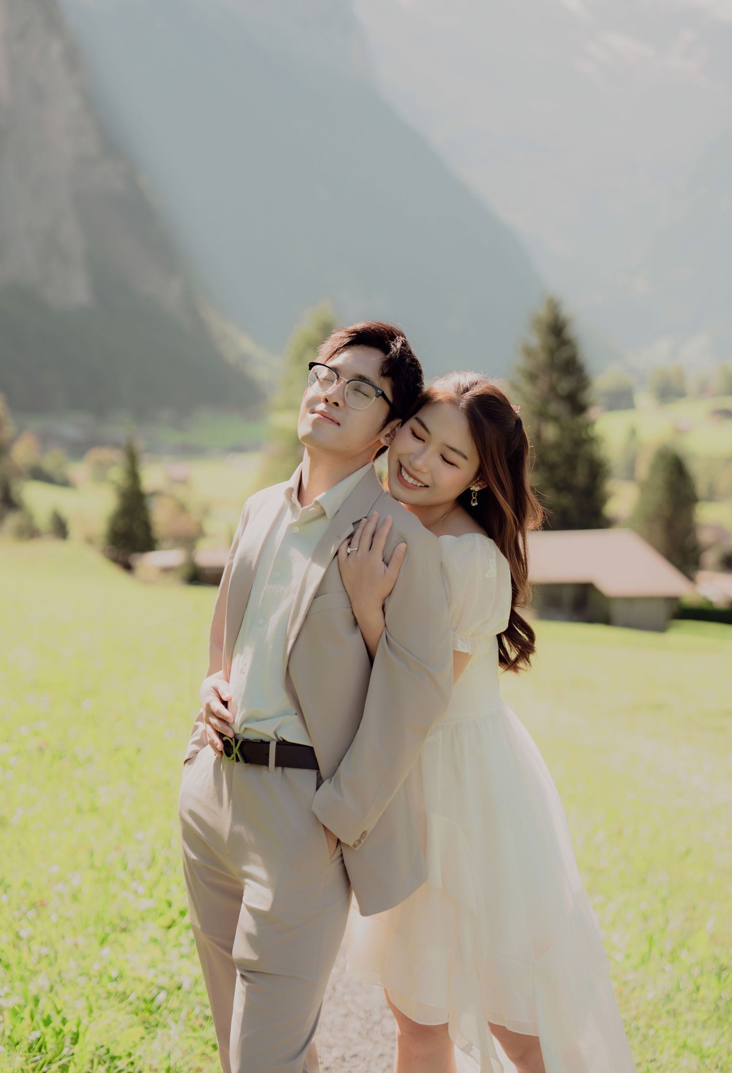 Bride hugging groom from behind as they smile on a sunny green meadow with Alpine mountains in background.