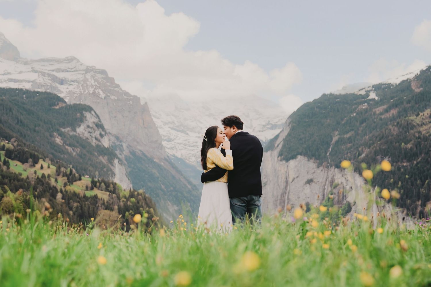 Couple sharing a romantic kiss in a lush alpine meadow with yellow wildflowers and snow-capped mountains in Switzerland.