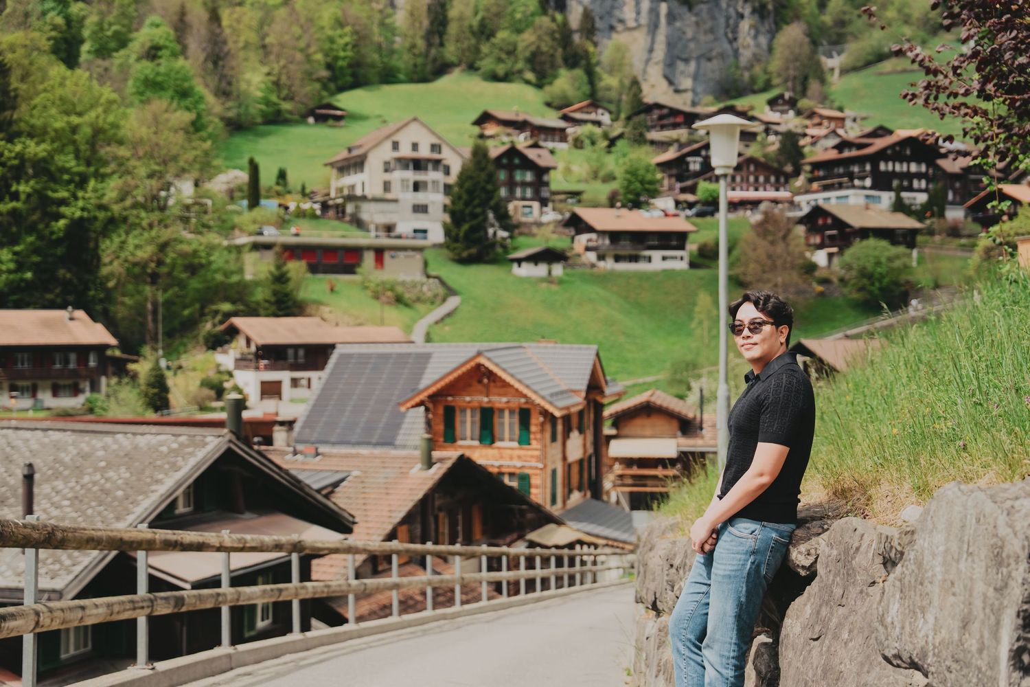 Man in black shirt and jeans posing on road with scenic Swiss chalet village on hillside behind.