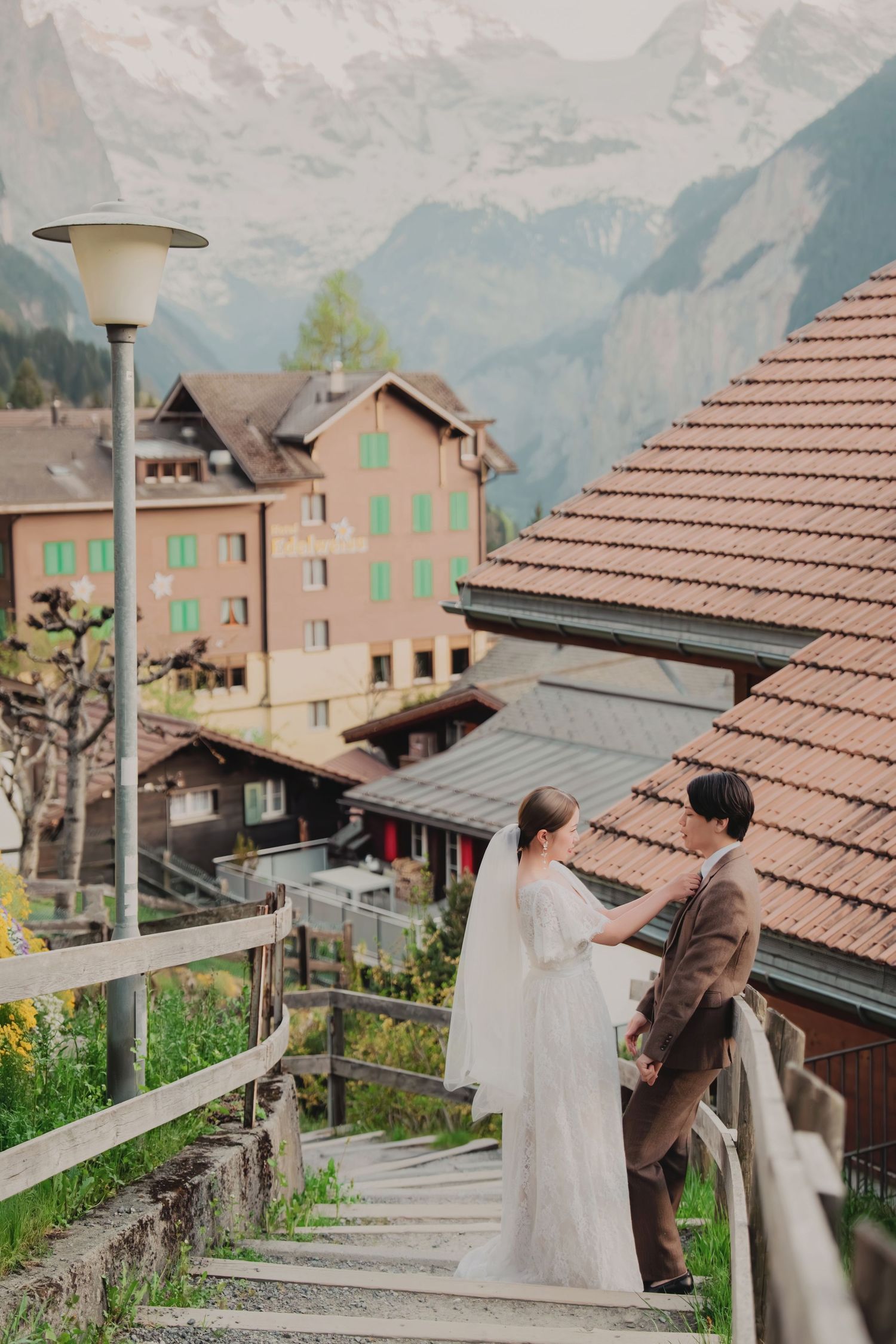 Wedding couple holding hands on scenic staircase overlooking a charming Swiss Alpine village with mountain backdrop.