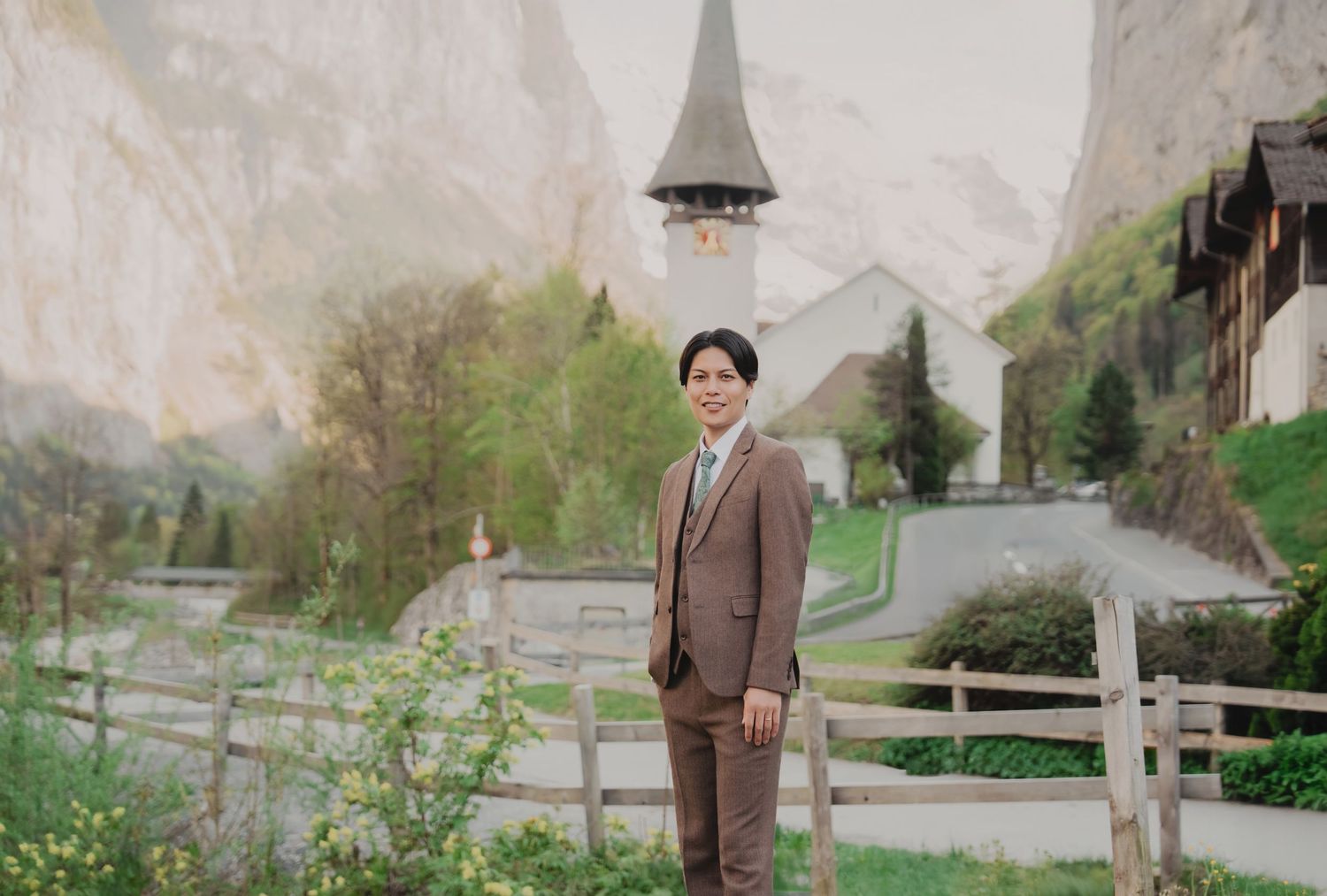 Smiling groom in brown suit poses before a scenic Swiss alpine village church.