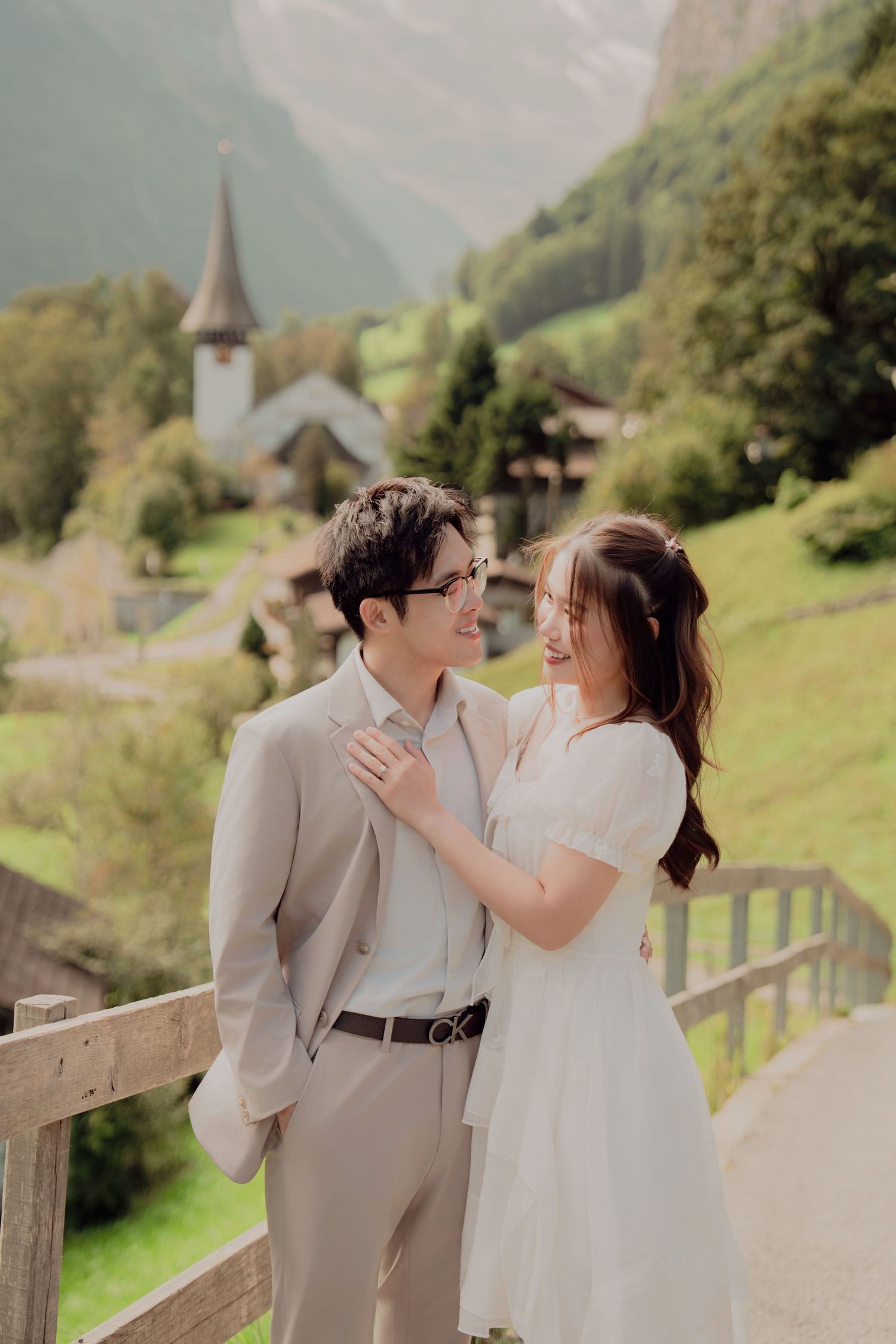 Couple gazing at each other on wooden bridge with Swiss village church and green mountains behind.