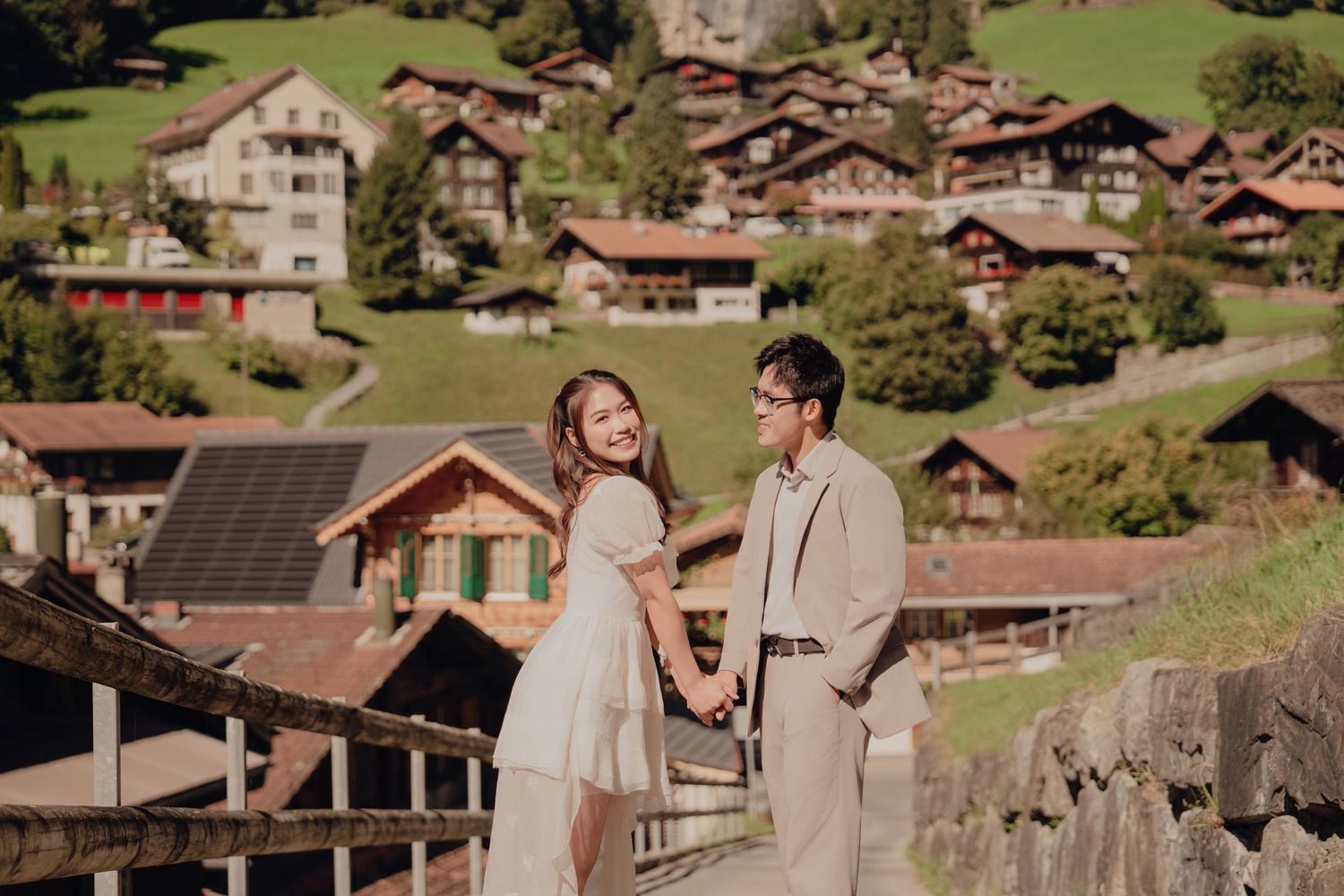 Couple holding hands smiling back at camera with Swiss chalet village on a hillside.