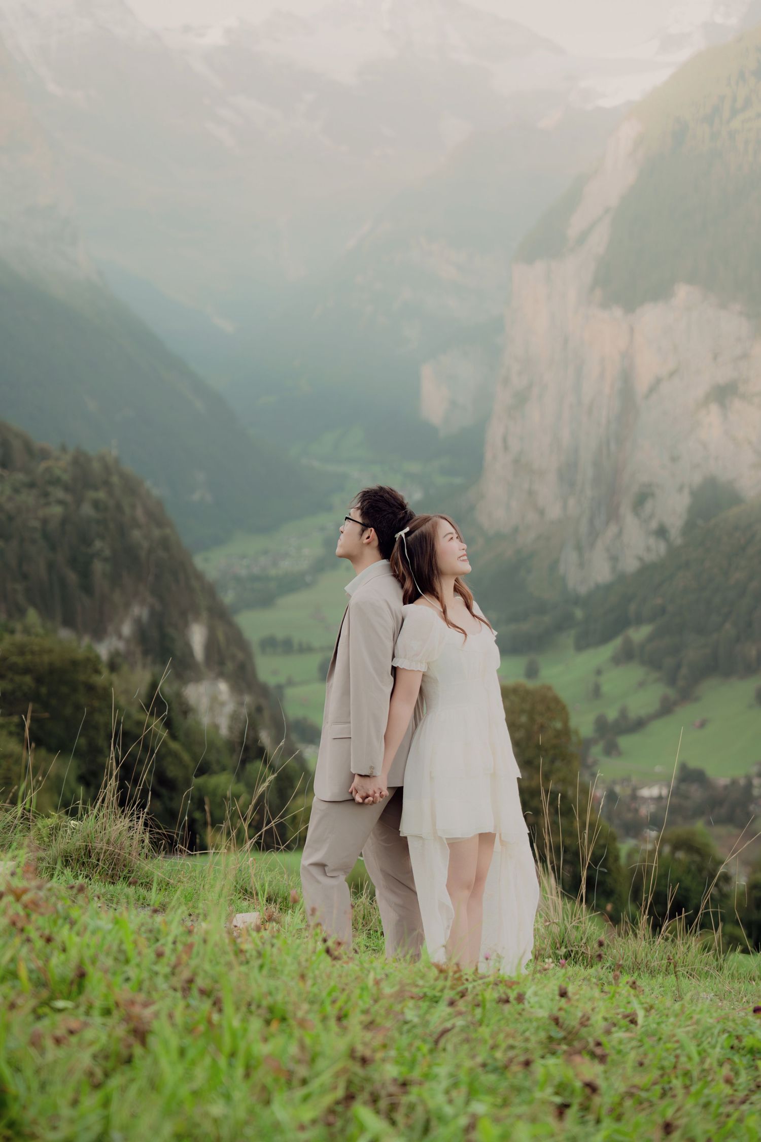 Bride and groom standing back-to-back holding hands on a hillside overlooking a breathtaking Alpine valley.