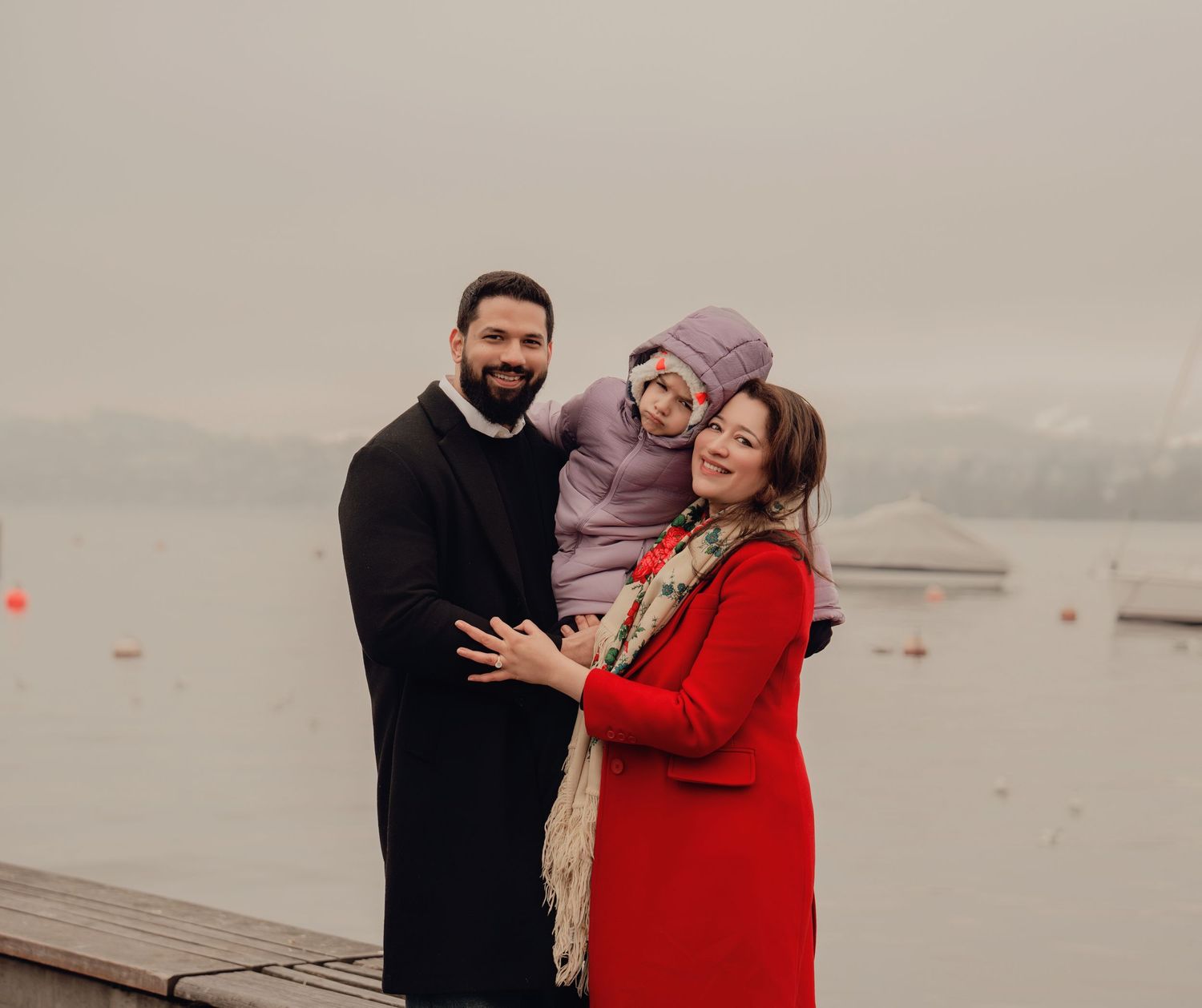 Family portrait by a foggy lake, father and mother in red coat holding their young child warmly.