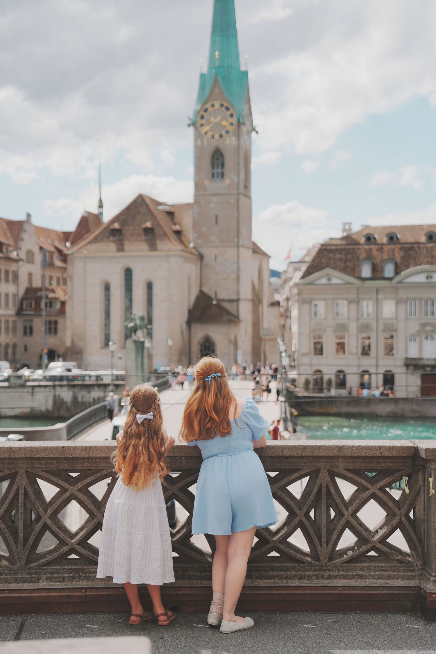 A woman and young girl in blue and white dresses view Fraumunster church from a bridge in Zurich.