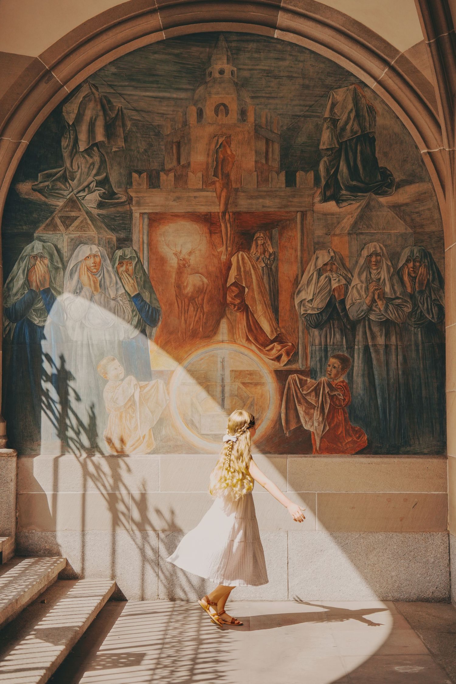 Girl in white dress walking past large religious fresco mural in sunlit arched corridor.
