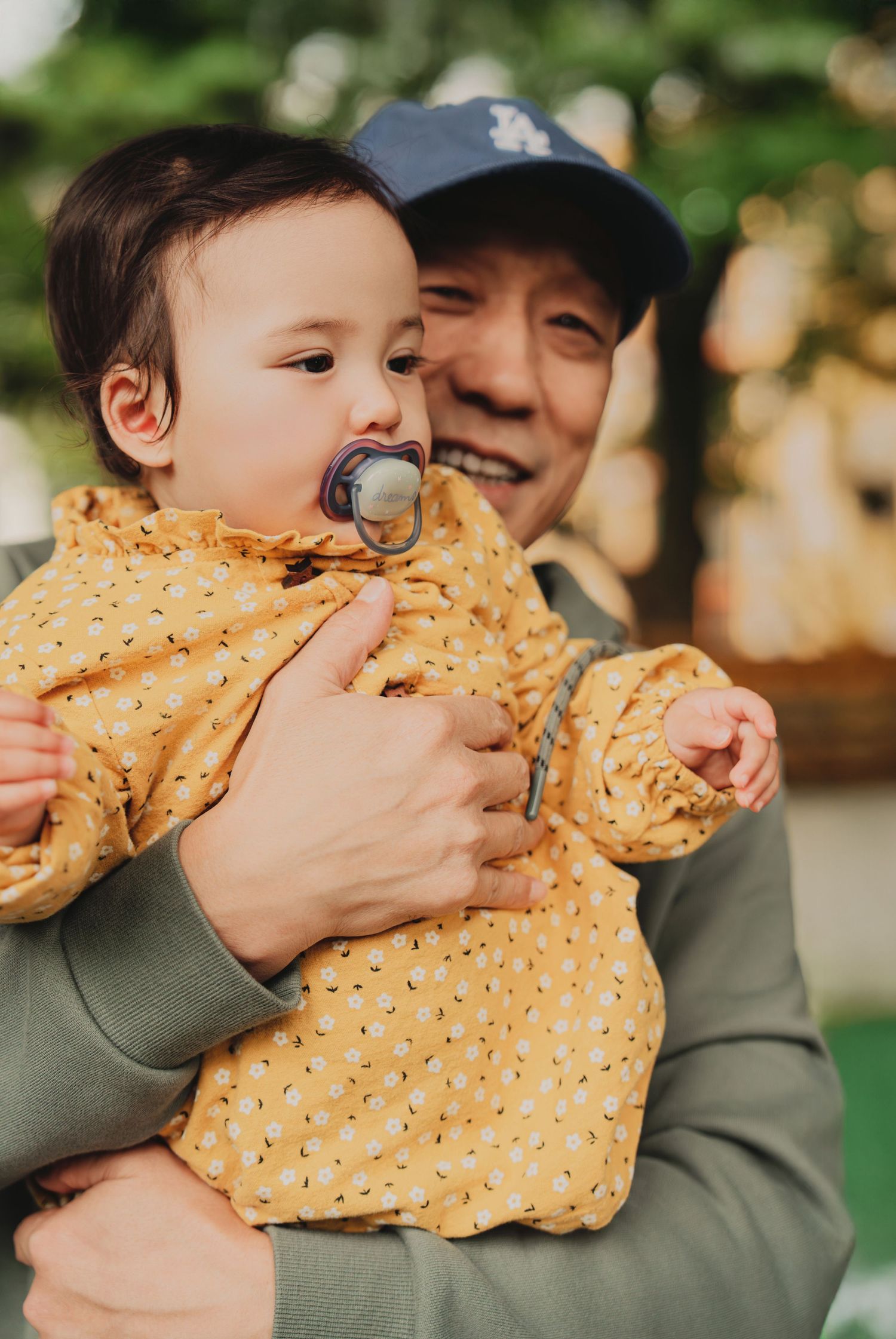 Smiling grandfather holding a baby girl in a yellow floral outfit with a pacifier outdoors.