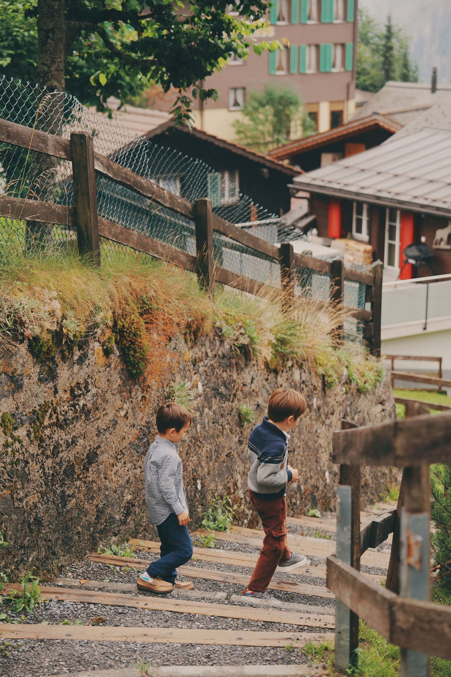 Two young boys walk up wooden stairs along a lush hillside in a picturesque alpine village.