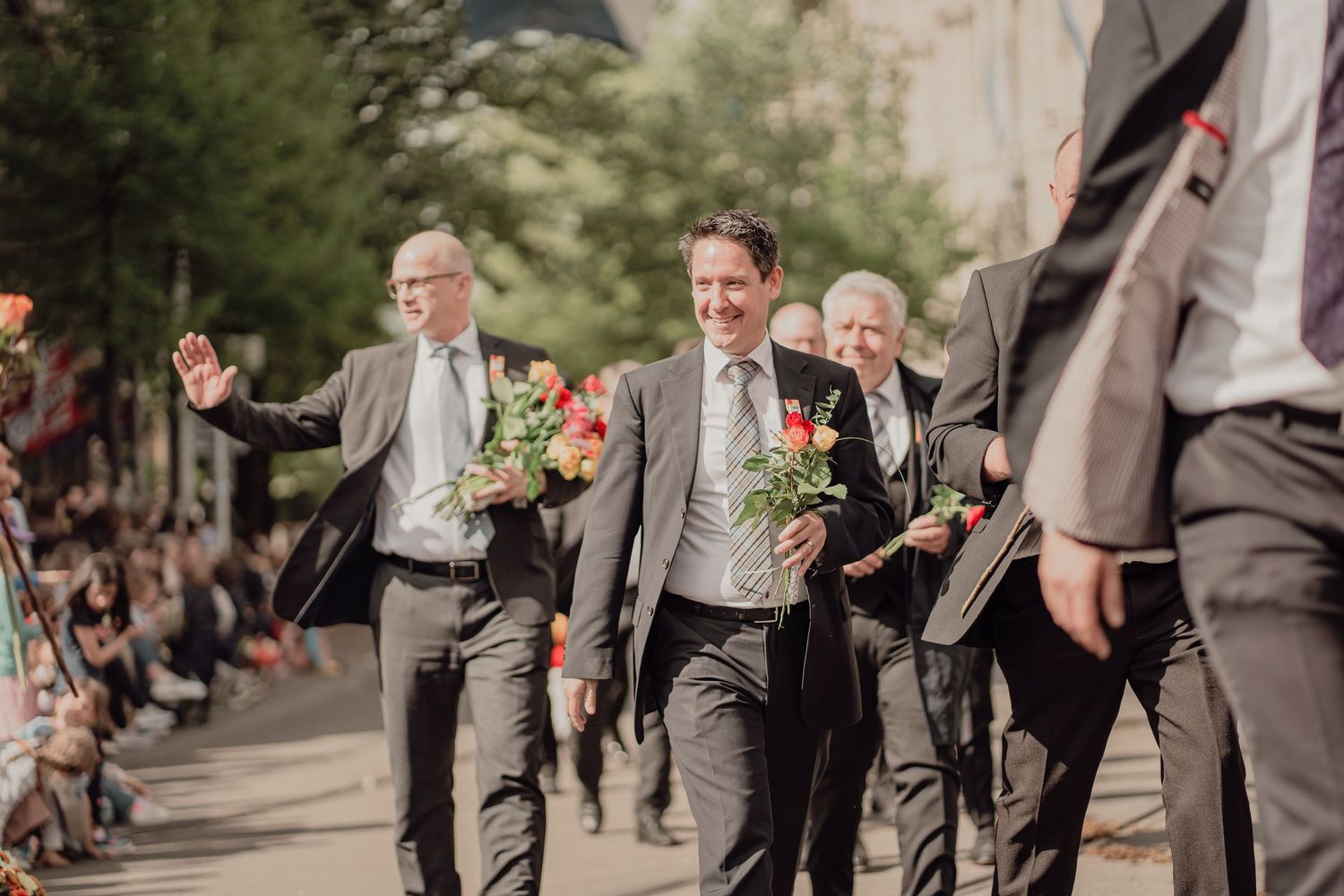 Smiling men in gray suits walk in a parade, waving and carrying colorful bouquets of roses.