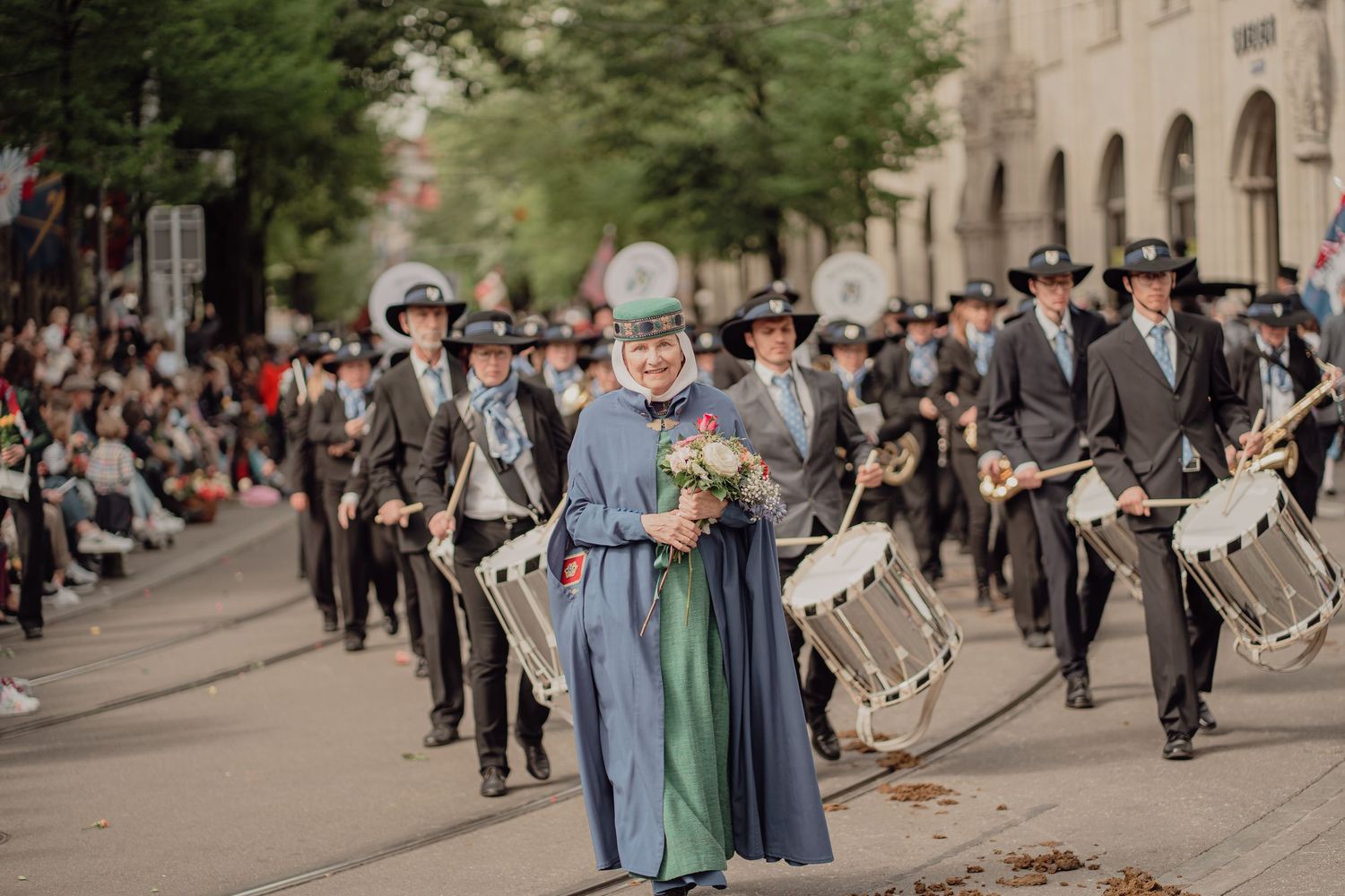 A woman in a blue dress leads a marching band with drummers in uniform through a city street parade.