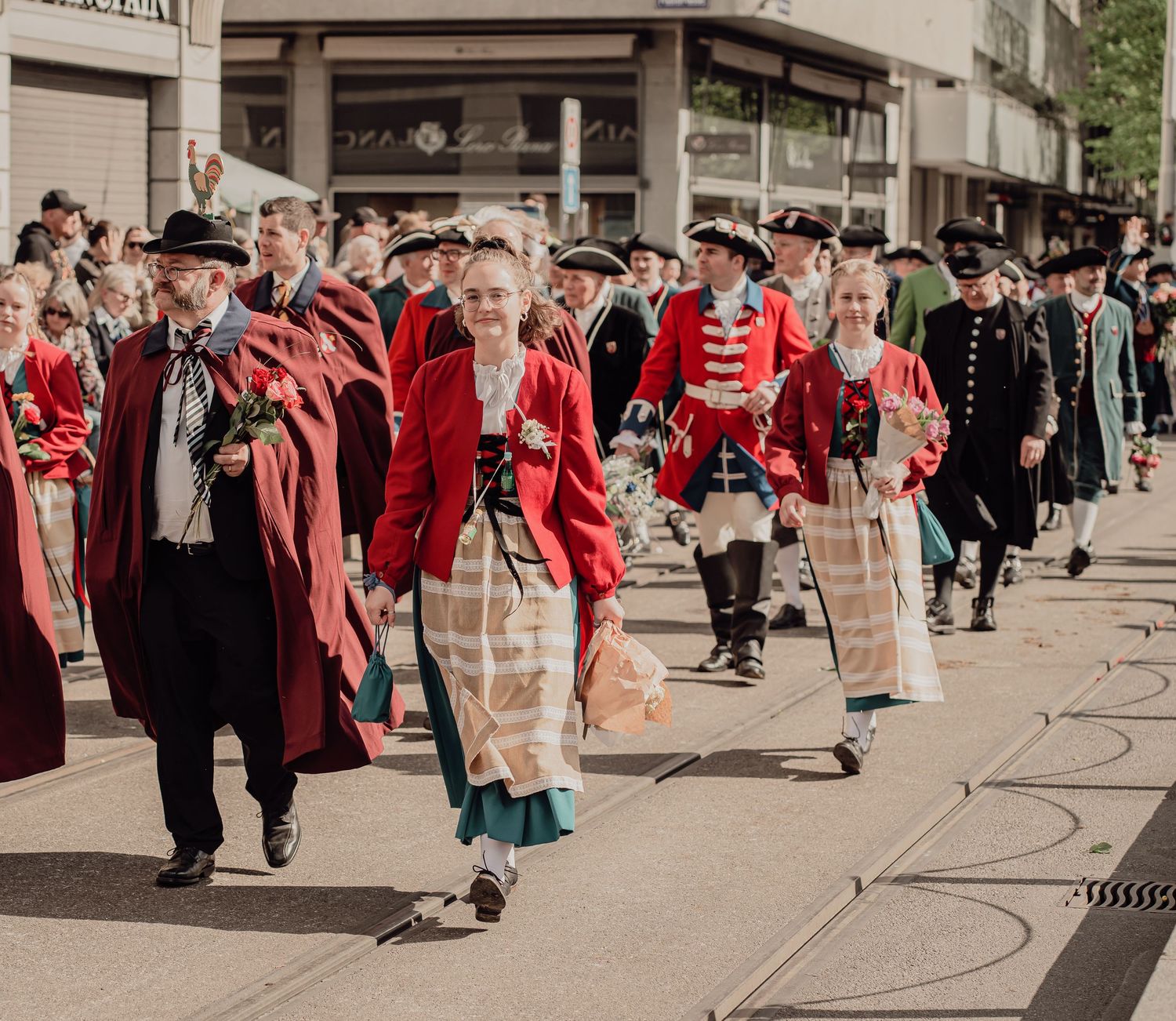Participants in red historical costumes and traditional attire march through city streets in a cultural parade.