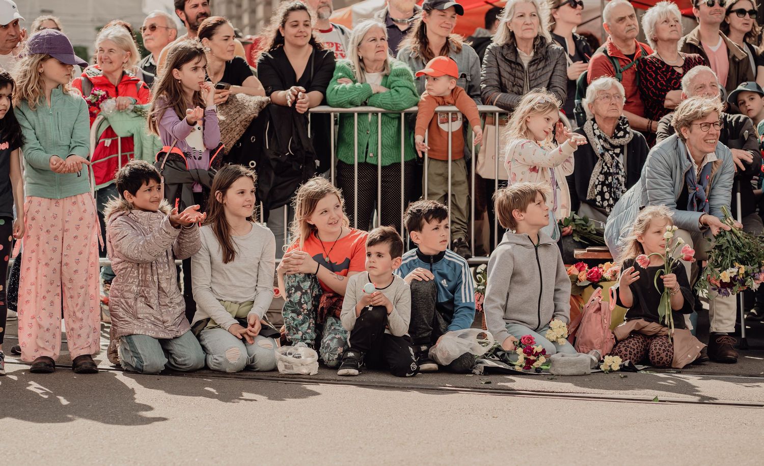 Children and adults watching a parade from the sidelines, sitting along a metal barrier outdoors.