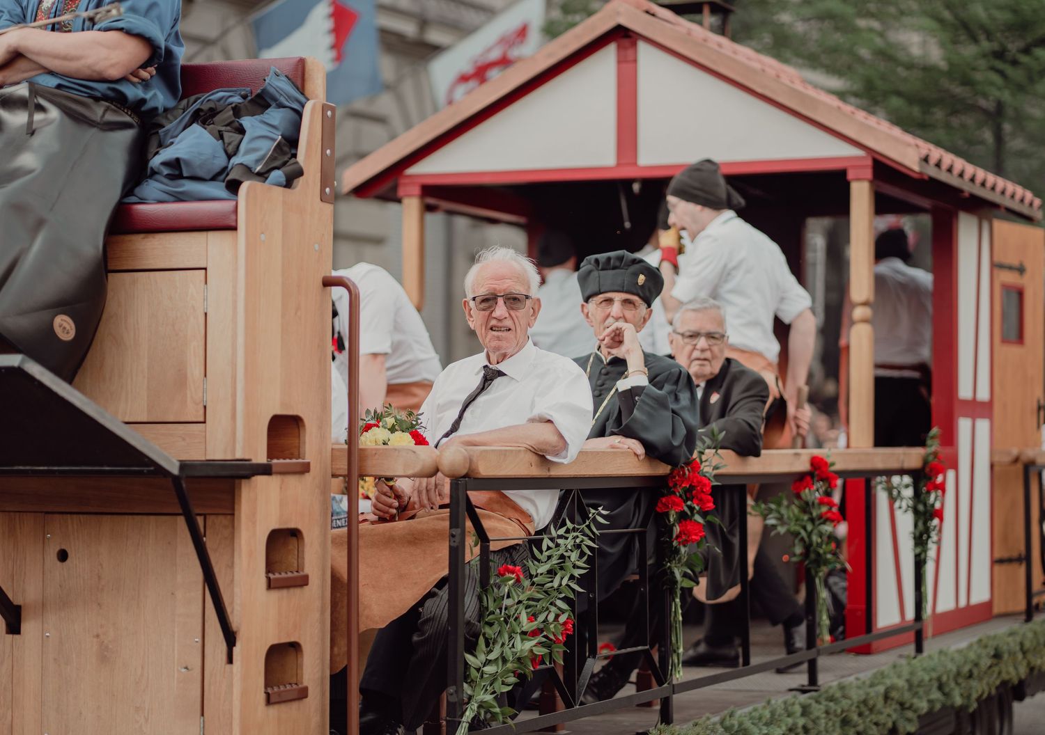 Elderly man sits on a wooden parade float with decorative red flowers during a traditional festival.