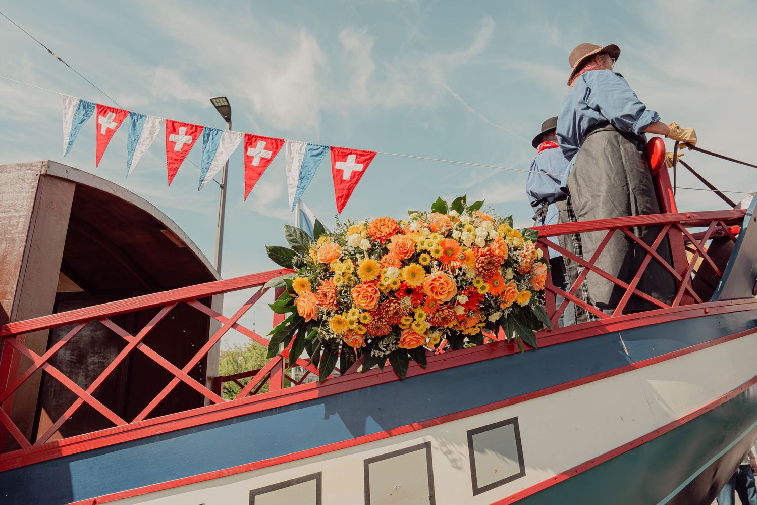 Person standing beside a large orange rose bouquet on a decorated boat with Swiss flag bunting at a festival.