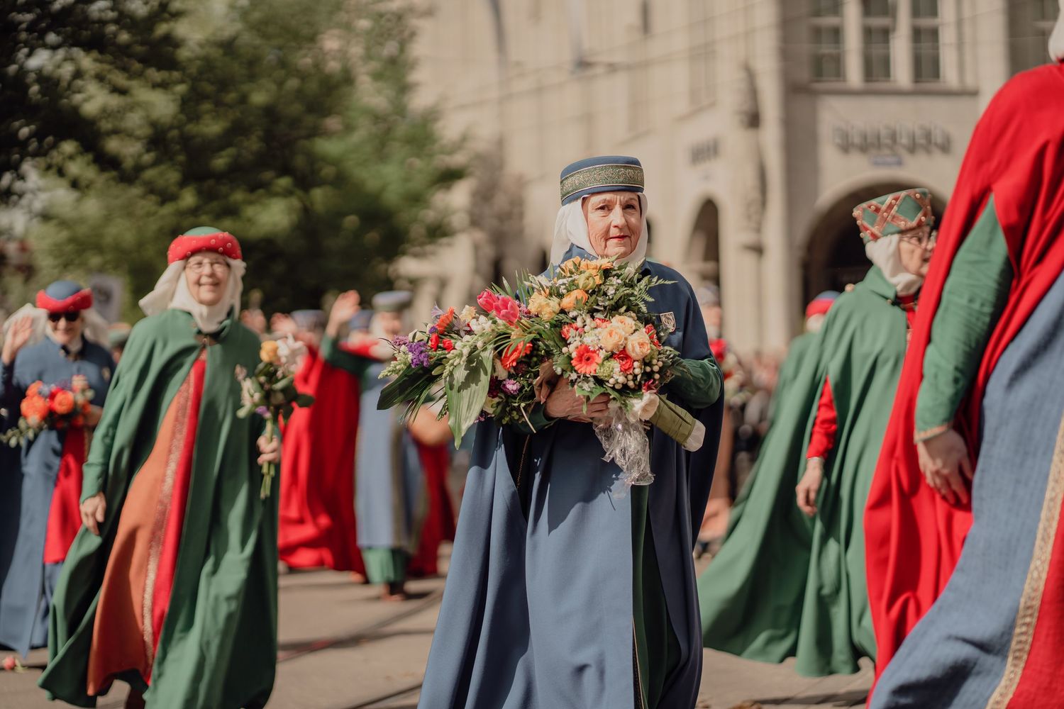 Woman in blue traditional robe carries large floral bouquet during a colorful medieval street parade.