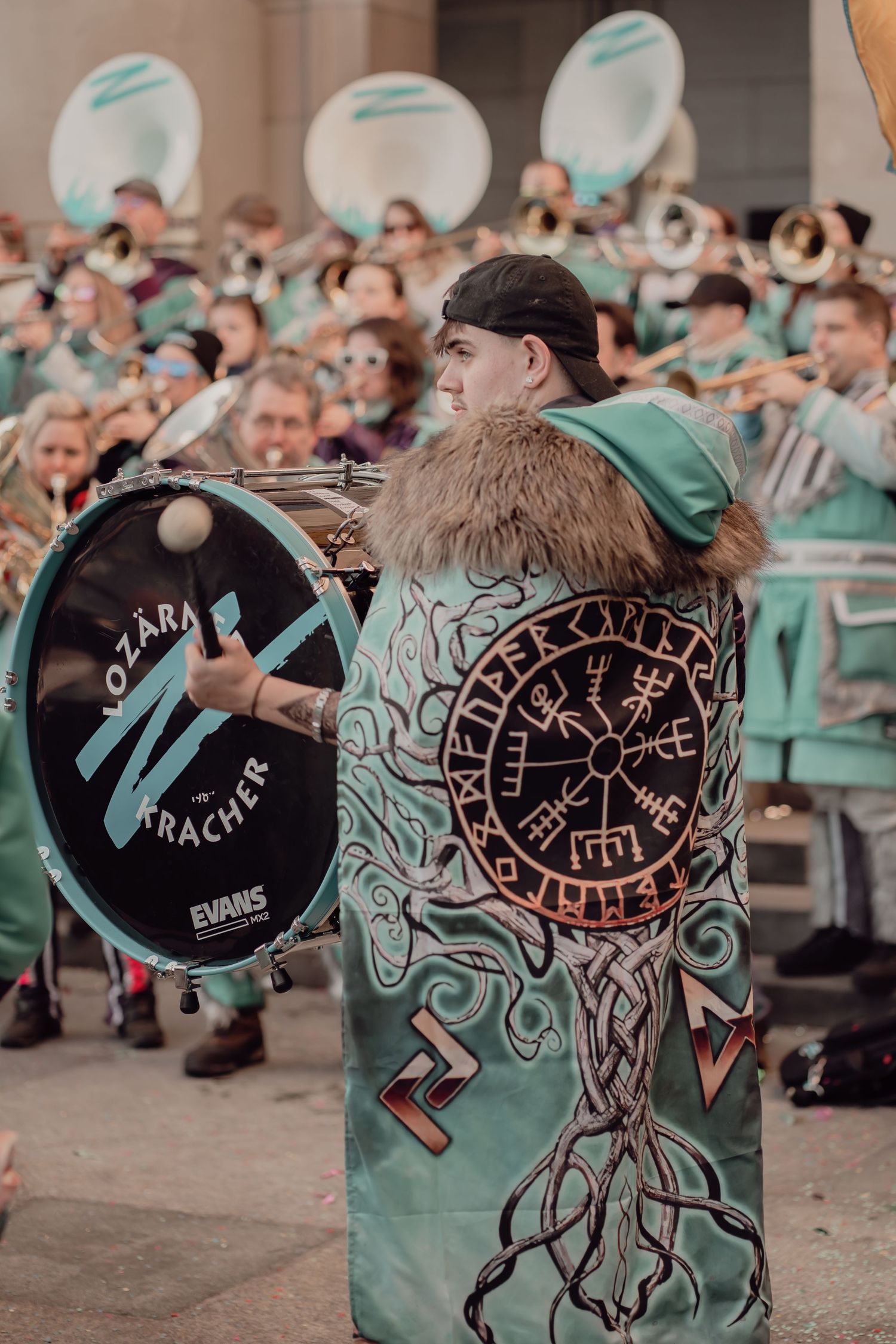 A drummer in a teal Viking-themed costume plays bass drum at an outdoor festival parade.