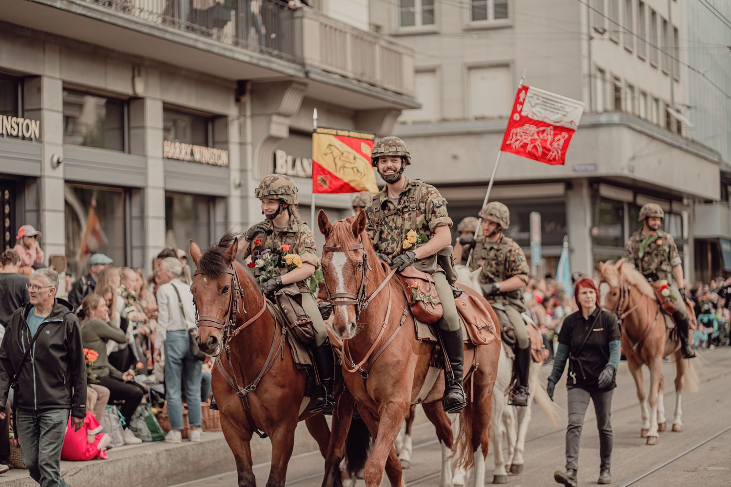 Soldiers on horseback carrying regional flags ride through a busy urban parade route.