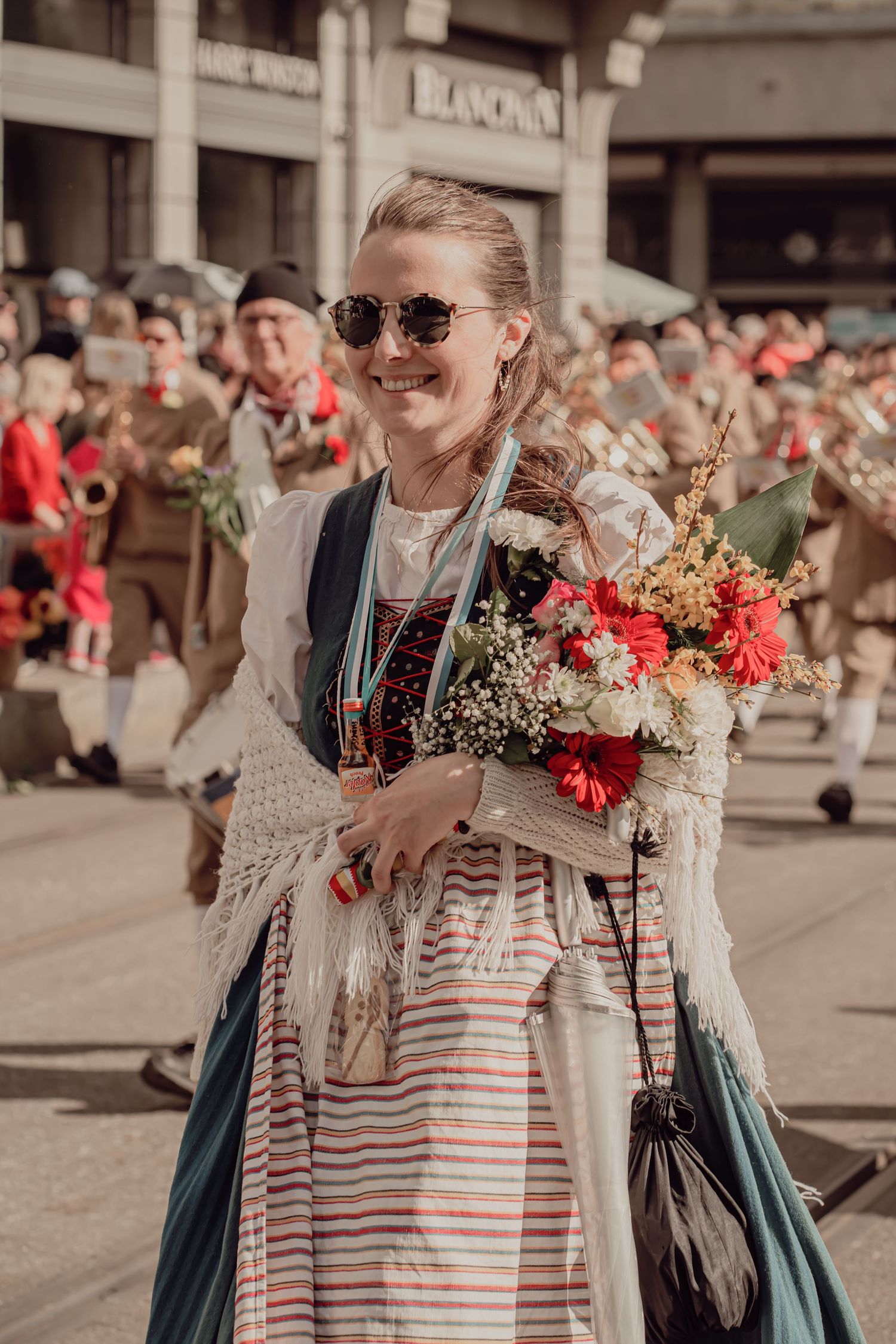 Smiling woman in traditional folk dress holds a bouquet of flowers at a festive parade.
