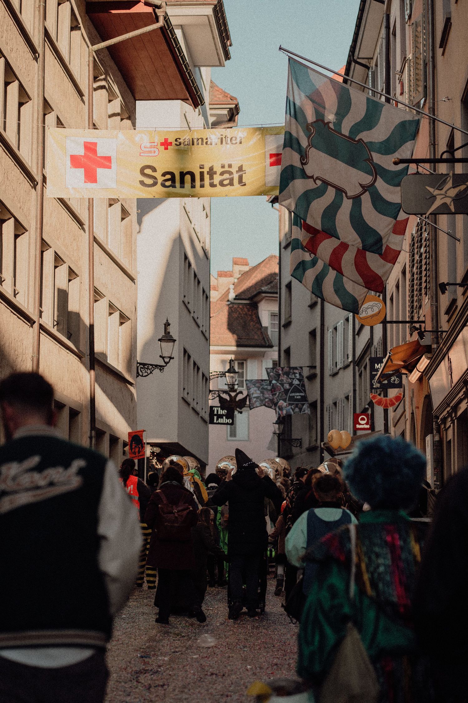 Crowded carnival street with Red Cross Sanitaet banner and colorful flags.