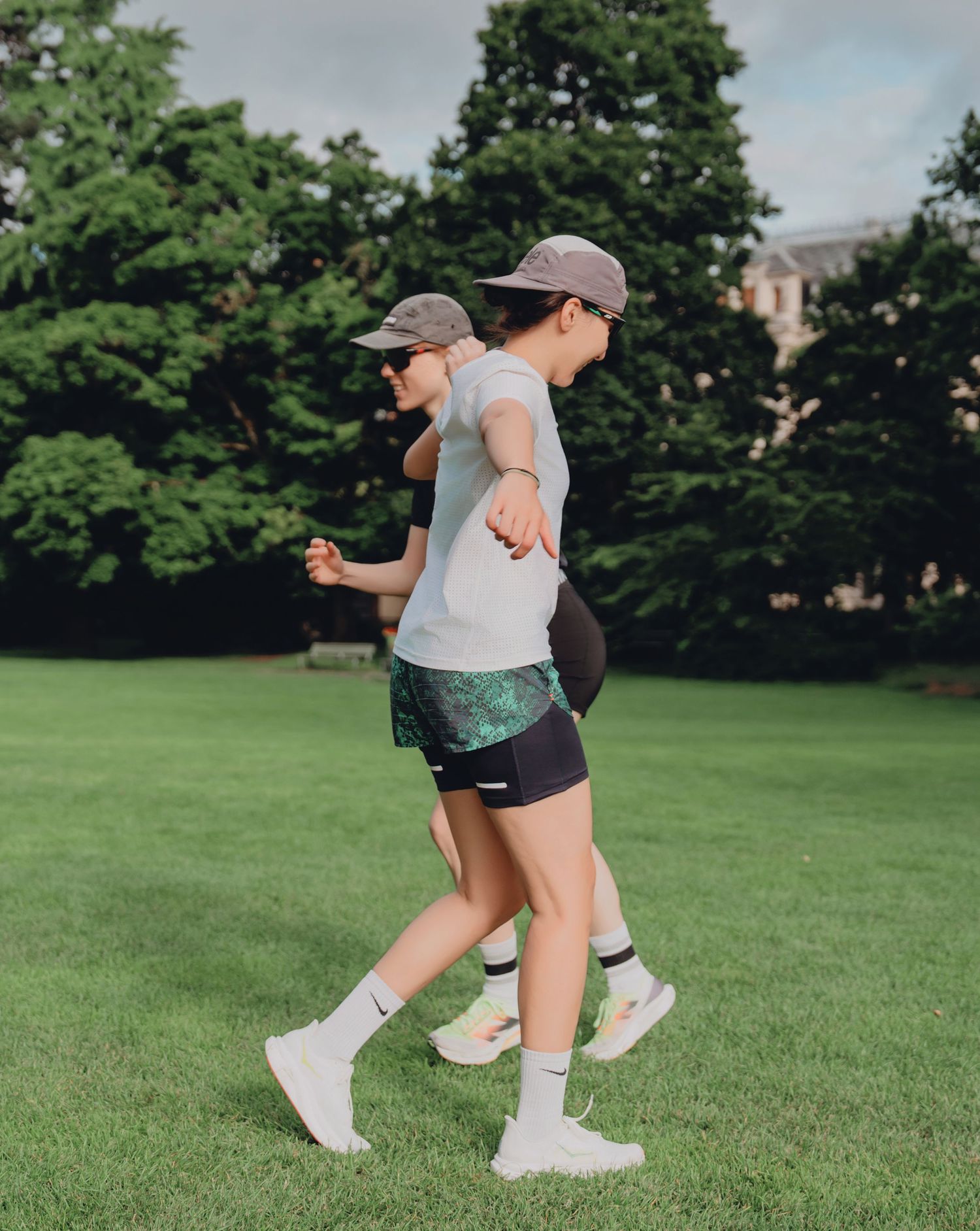 Two women in athletic shorts and caps walking across a green park lawn with historic building in background.