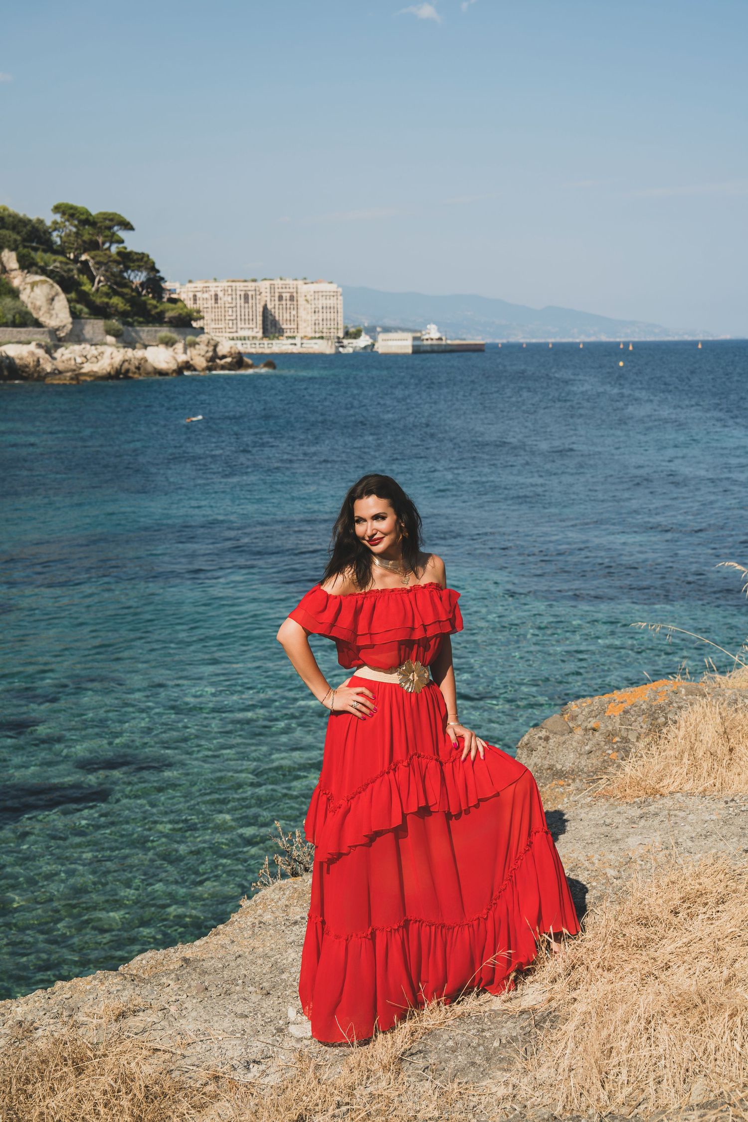 Smiling woman in flowing red off-shoulder dress standing on rocky Mediterranean coastline.