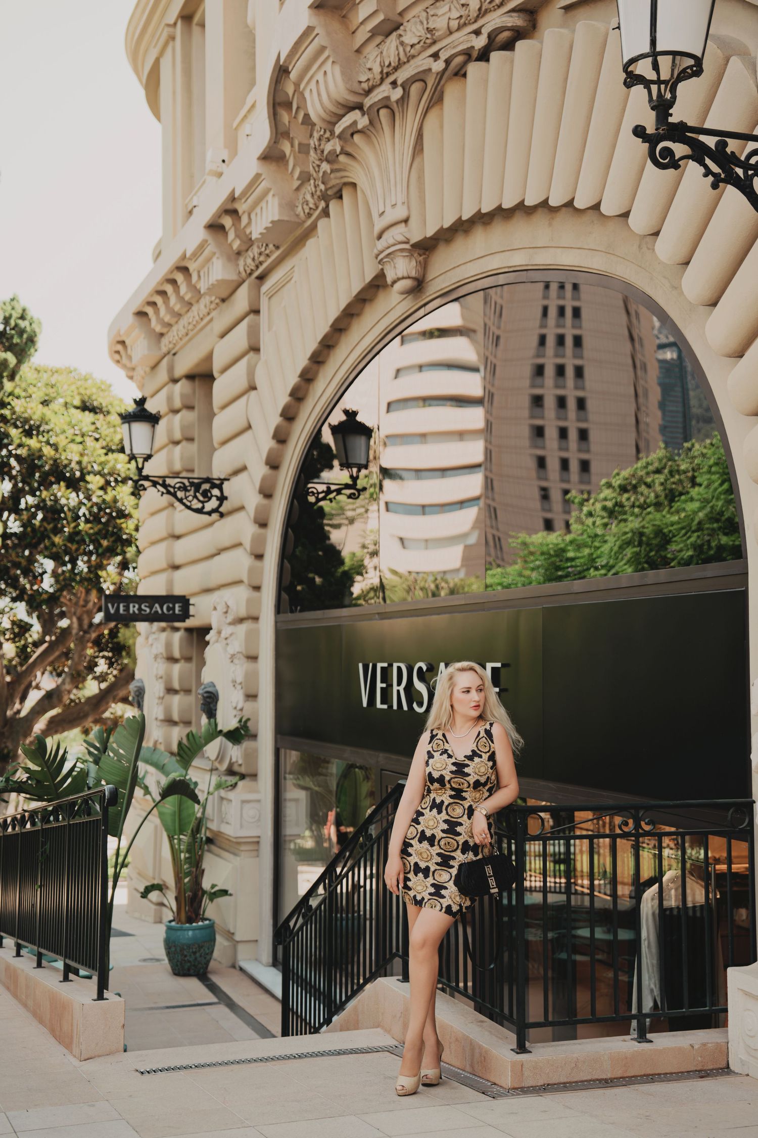 Elegant blonde in gold print mini dress standing under arch outside Versace boutique Monaco.