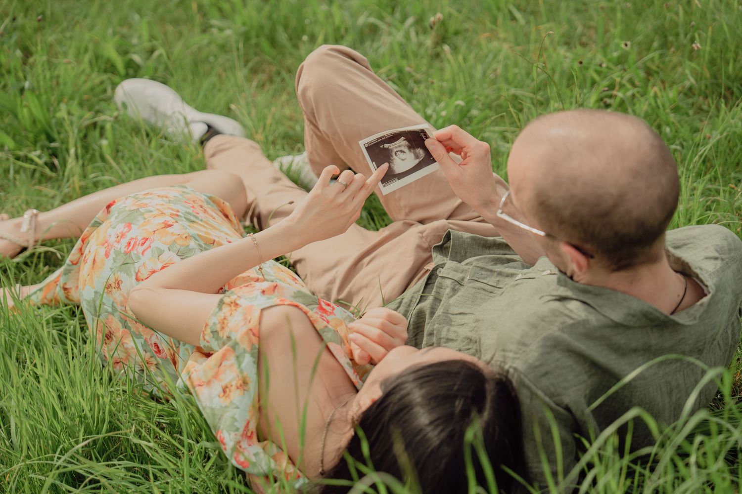 Couple lying in lush grass from behind, holding up a baby ultrasound during a maternity session.