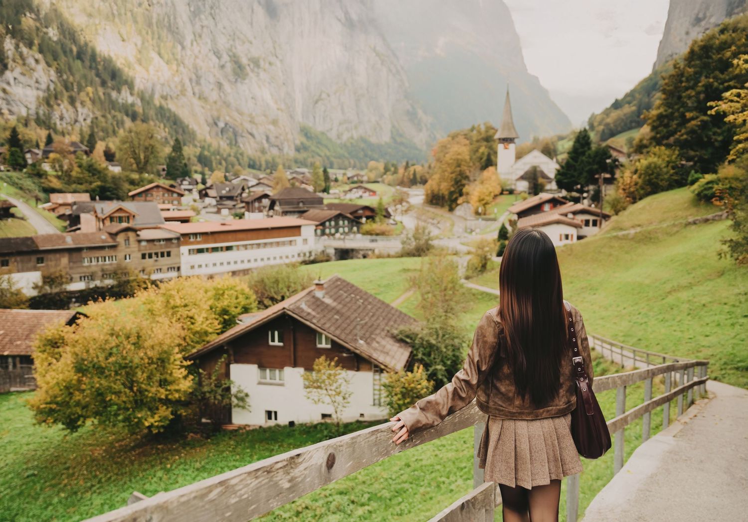 Woman in brown outfit stands by wooden fence gazing at scenic Swiss village nestled in alpine valley.