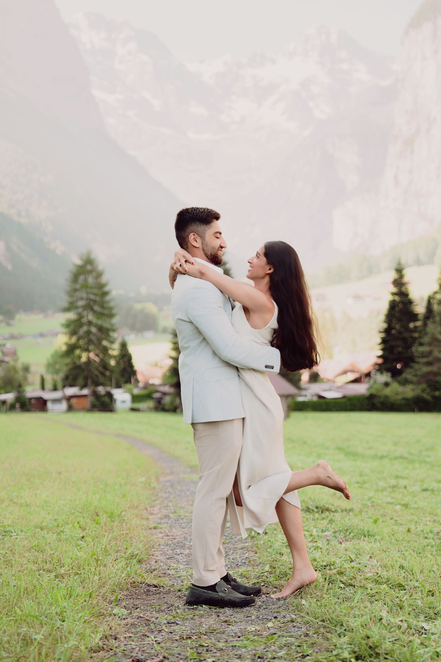 Joyful couple spinning and embracing in a green meadow with dramatic Swiss mountain backdrop.