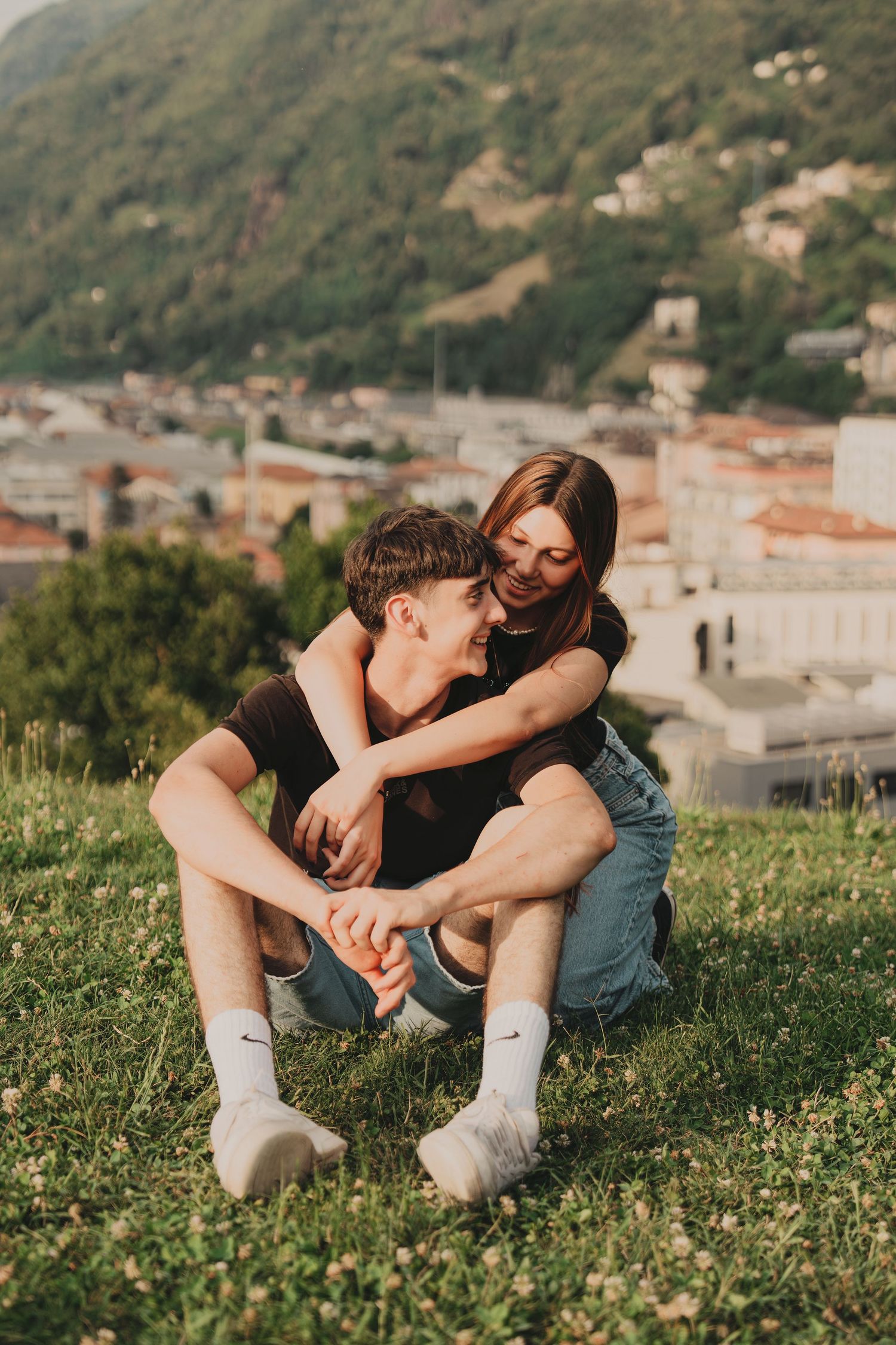 A young couple sitting on a grassy hill, embracing tenderly with a scenic town and mountains in the background.