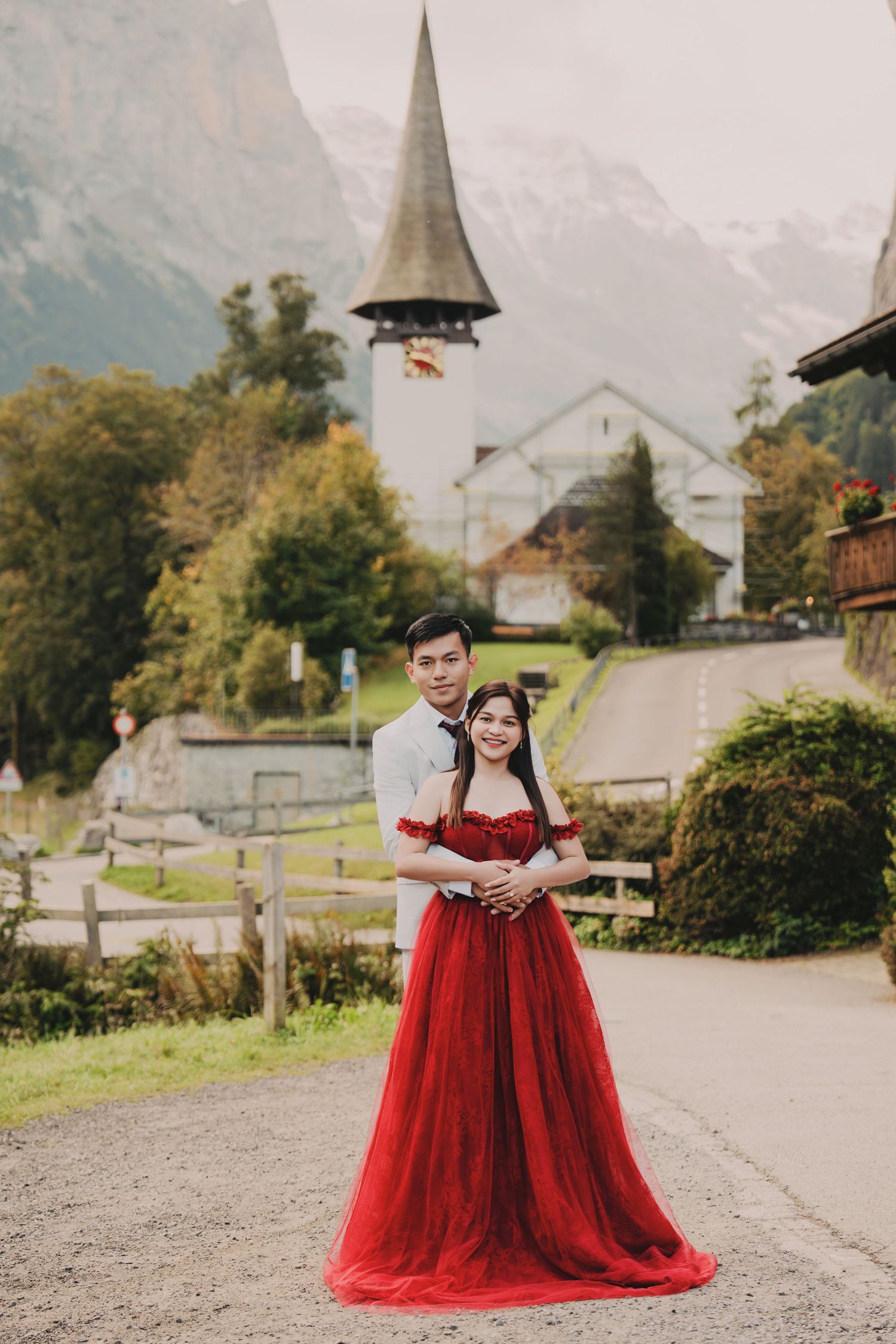 Couple embracing in front of Swiss village church, woman in red gown, man in white suit.