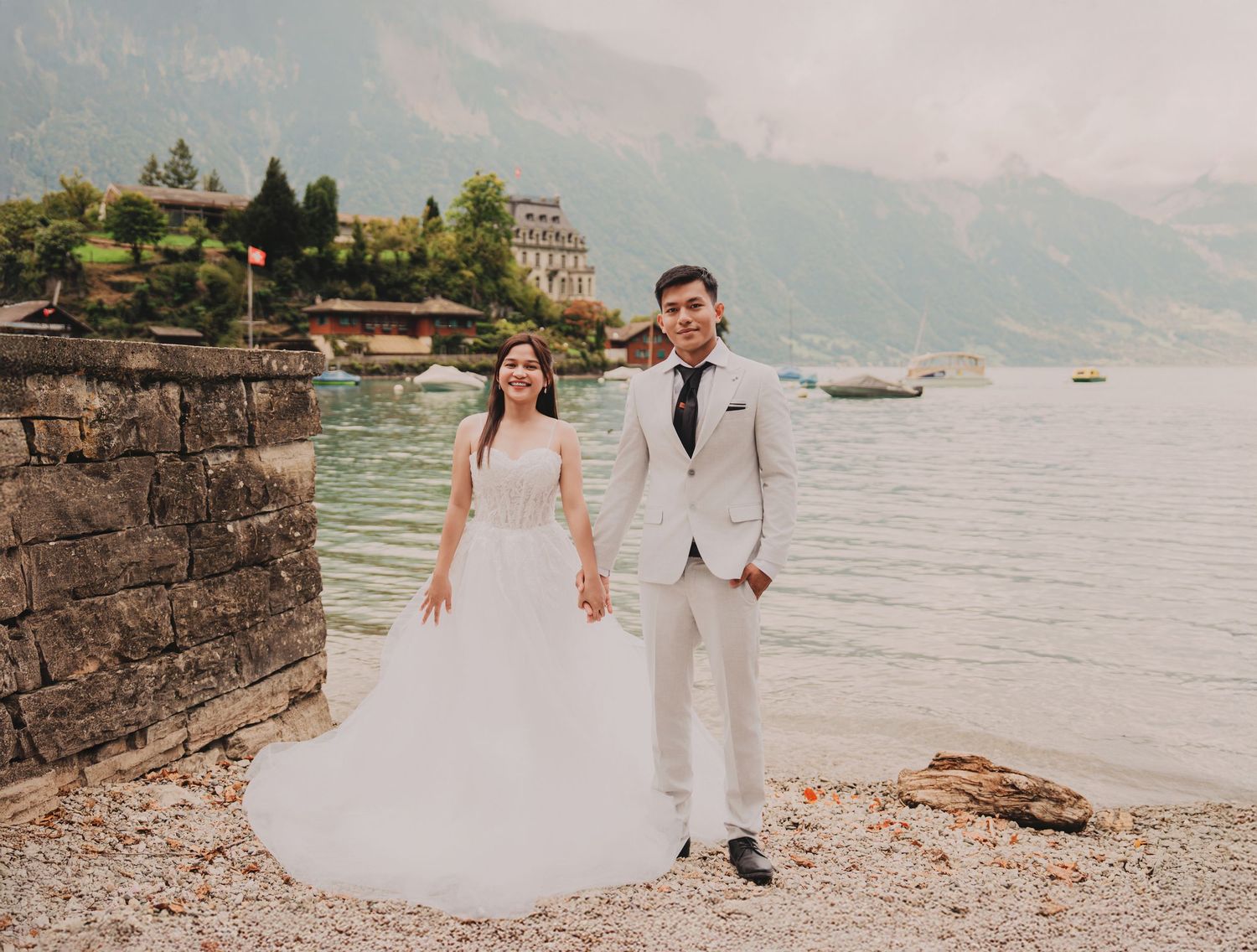 Bride and groom posing confidently by a scenic alpine lake with a grand historic building and mountains.