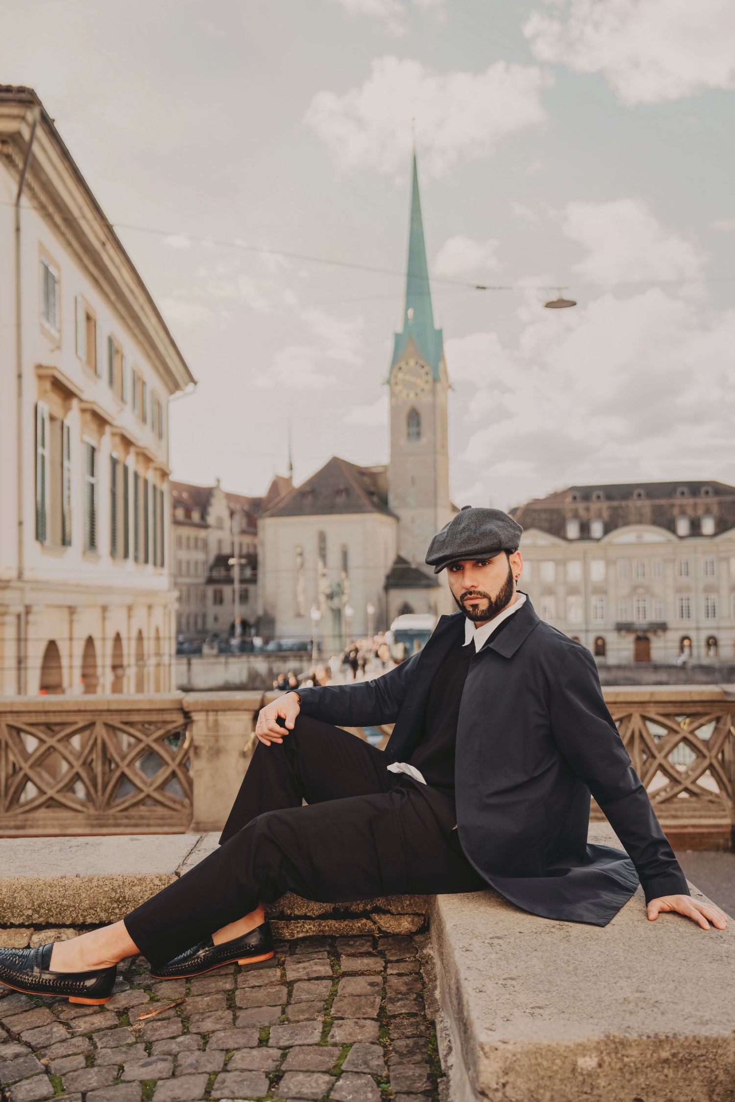 Stylish man in black coat and newsboy cap sits casually on stone wall with Zurich Fraumunster church behind.