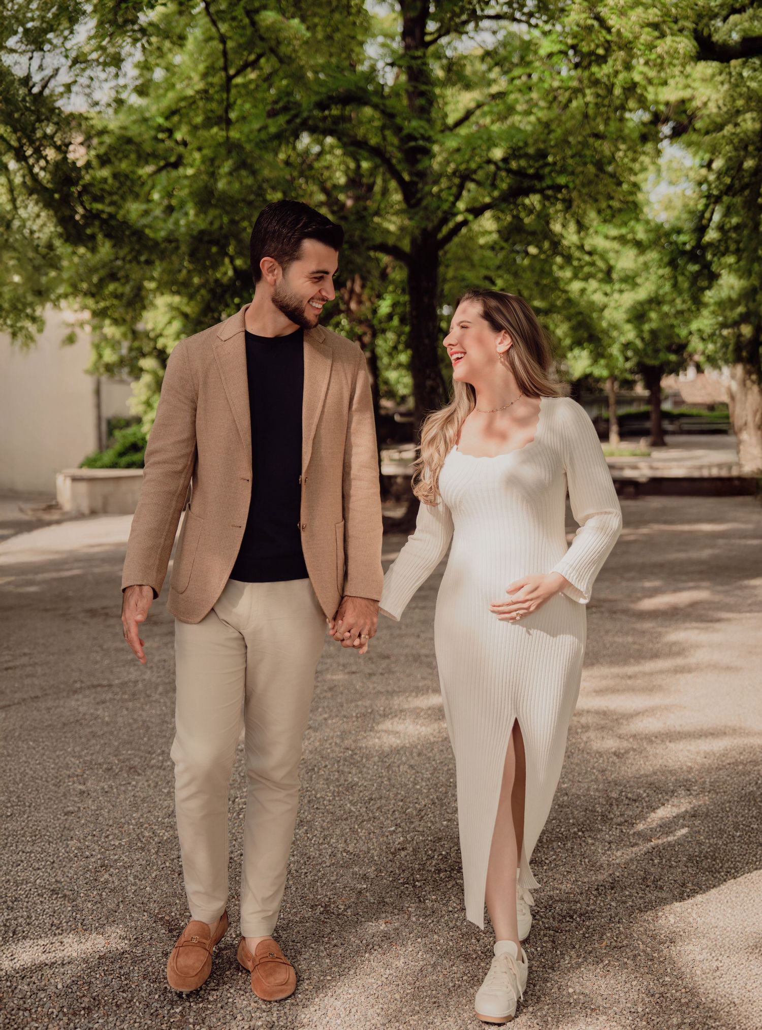 Expecting couple walking hand in hand outdoors, woman in white maternity dress cradling baby bump, man in tan blazer.