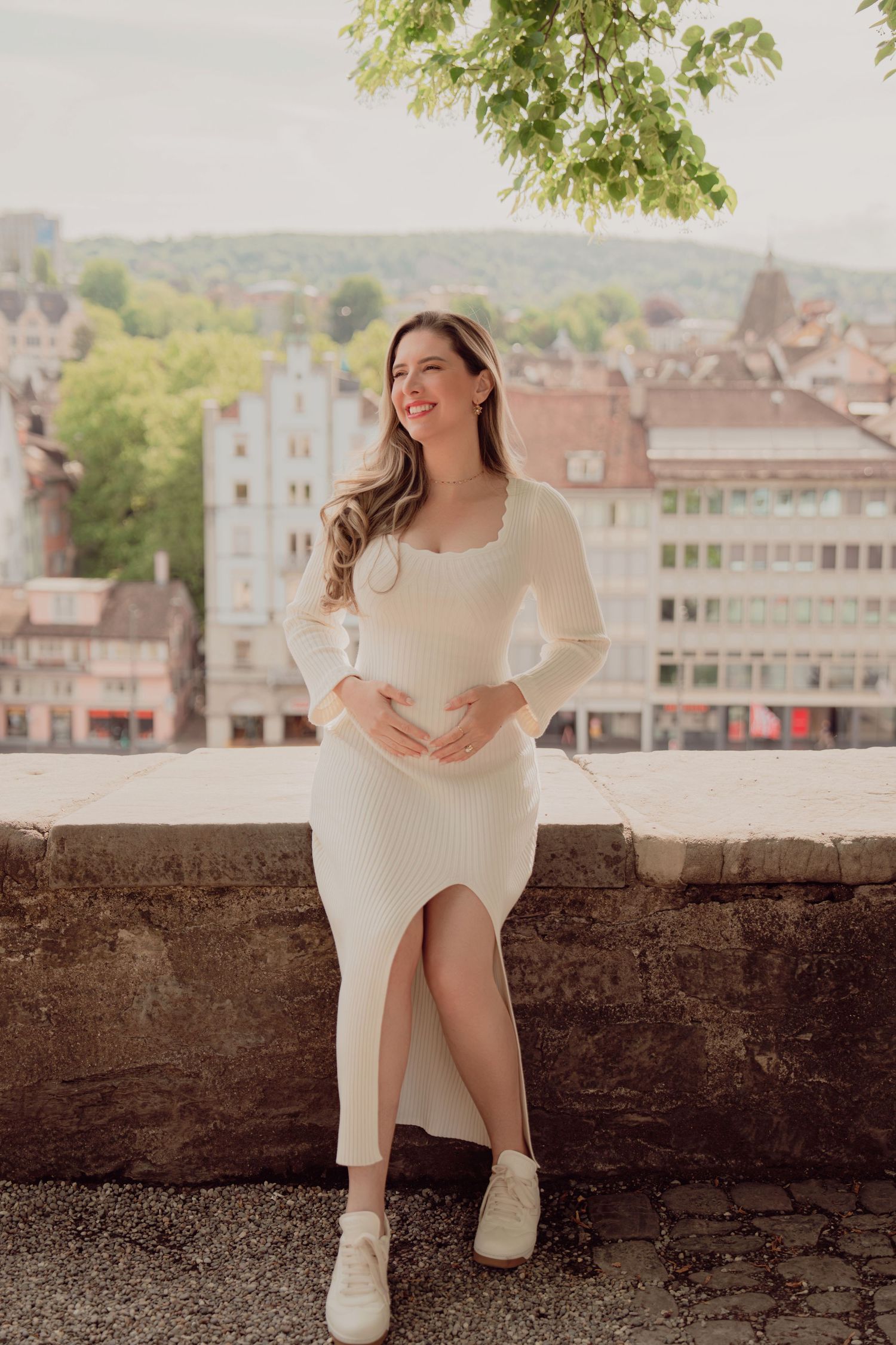 Smiling pregnant woman in cream ribbed dress sits on stone wall with Zurich city panorama behind her.