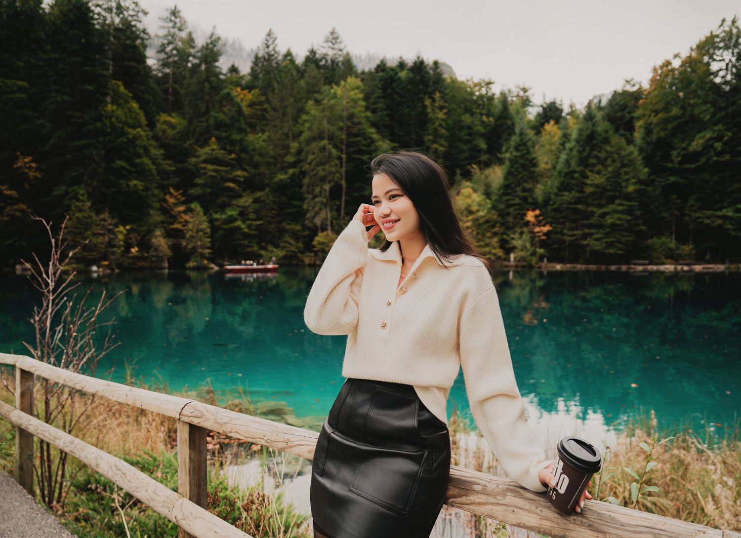 Smiling woman in cream polo sweater and black skirt holding coffee cup by turquoise mountain lake.