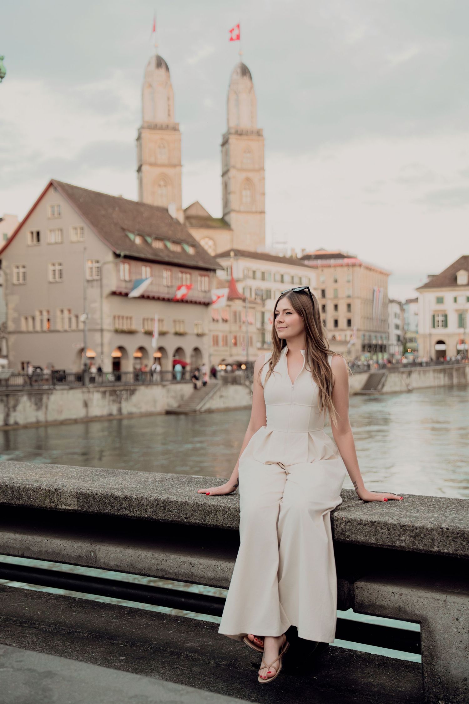 Elegant woman in beige wide-leg outfit sitting on bridge railing with Grossmunster cathedral backdrop.