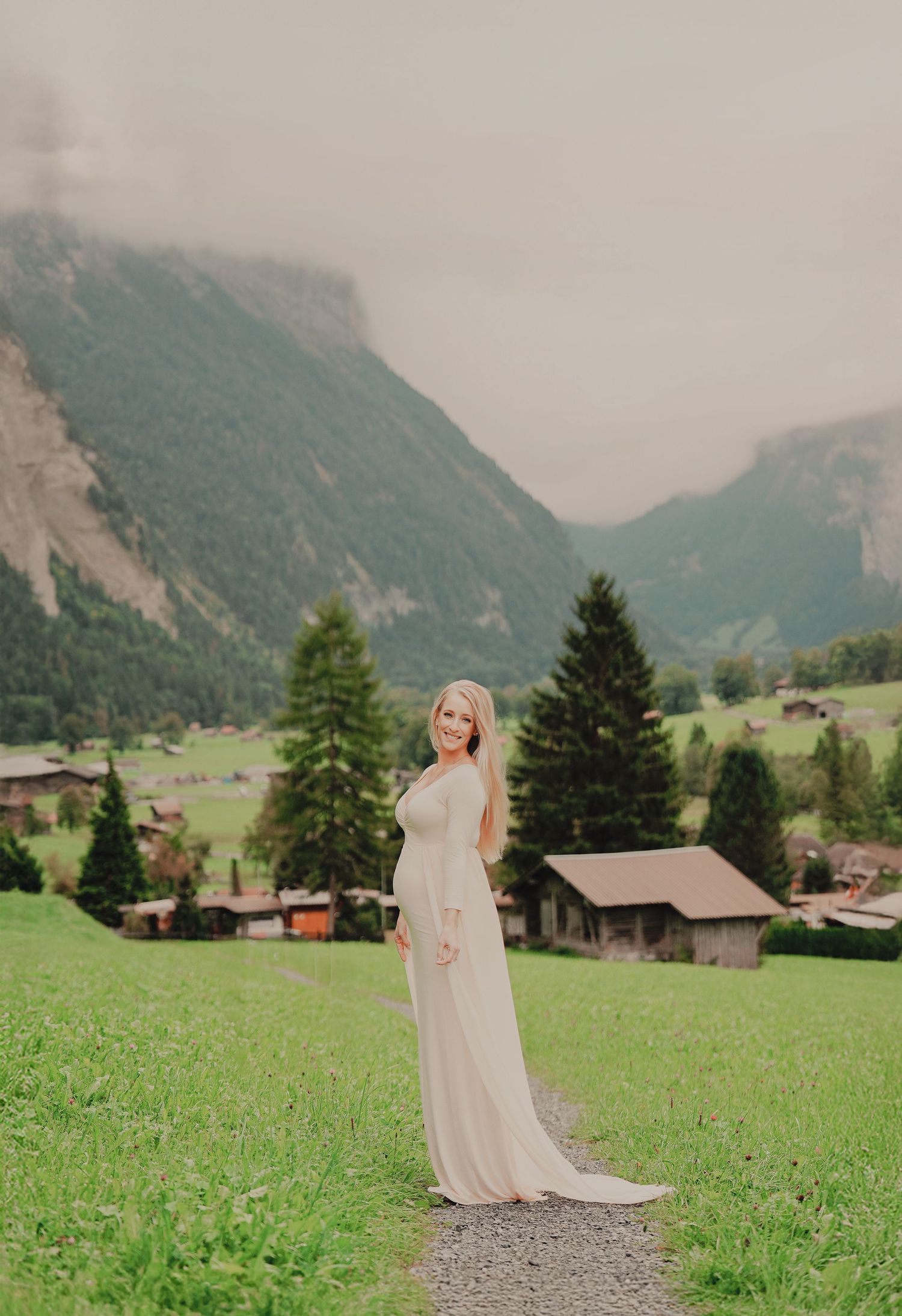Pregnant woman in flowing white gown glancing back while standing on a path with misty Swiss mountain scenery.