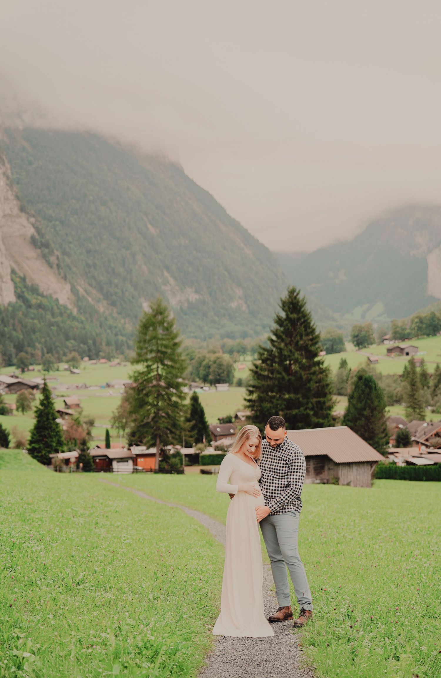 Expecting couple standing on gravel path in lush green Lauterbrunnen valley with misty mountains behind.