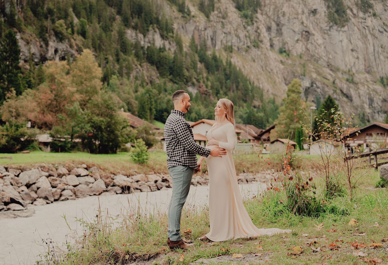 Expectant couple holding hands and smiling during outdoor maternity session in lush Swiss mountain village setting.