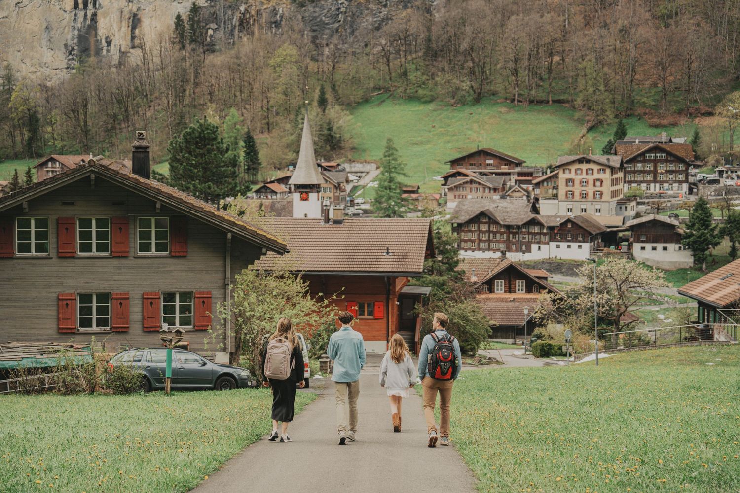Family walking toward a charming Swiss Alps village with traditional wooden chalets and green meadows.