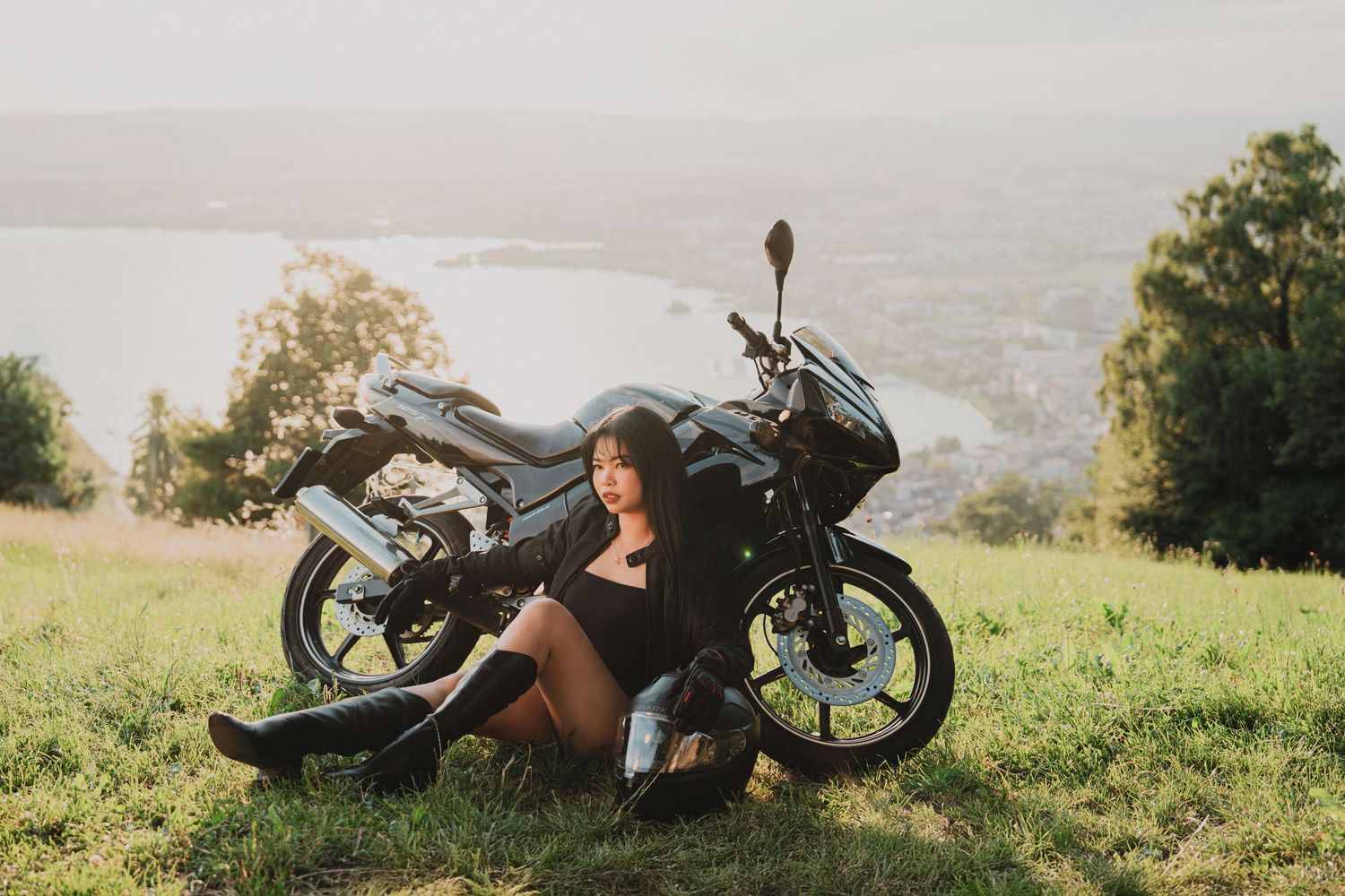 Young woman in black outfit sitting beside a black motorcycle on a sunny hillside overlooking a scenic lake.
