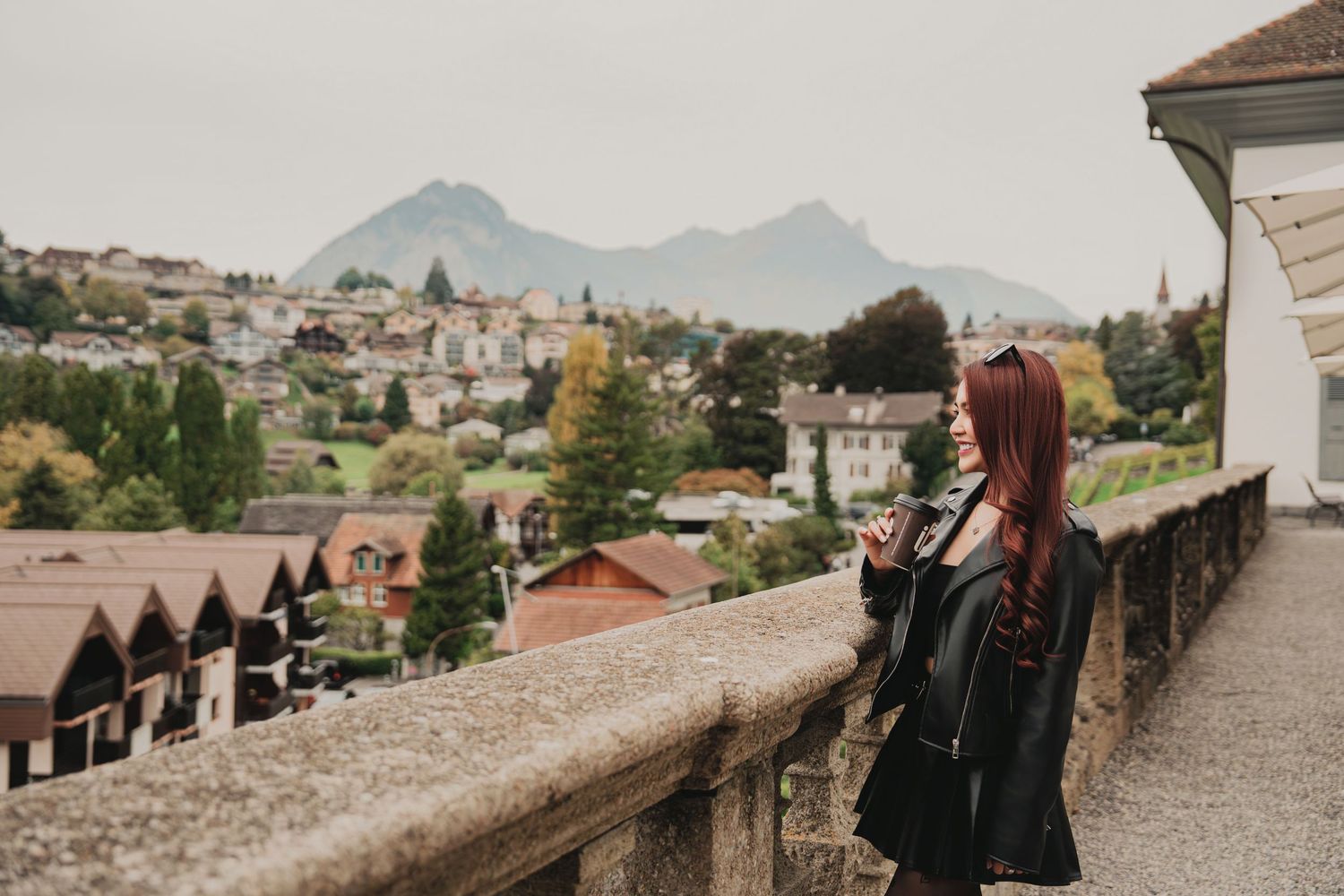 Red-haired woman in leather jacket standing on stone terrace overlooking a European mountain town in autumn.