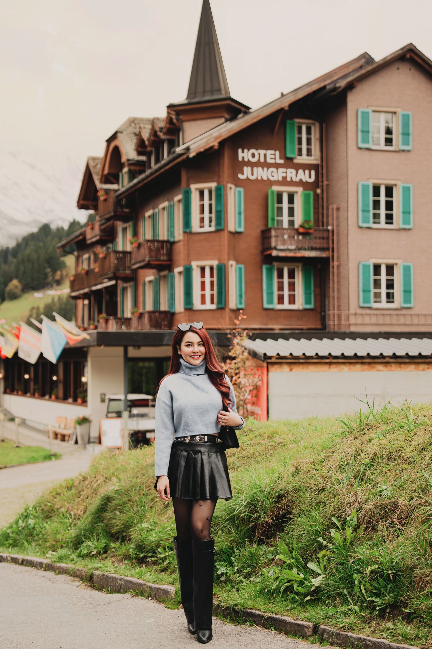 Woman in grey turtleneck and black leather skirt poses before Hotel Jungfrau in a Swiss Alpine village.