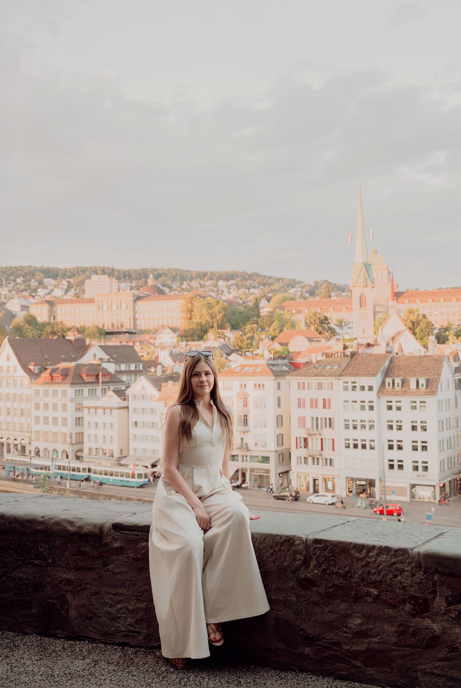 Woman in white jumpsuit sitting on stone wall with European cityscape and Gothic church spire behind her.