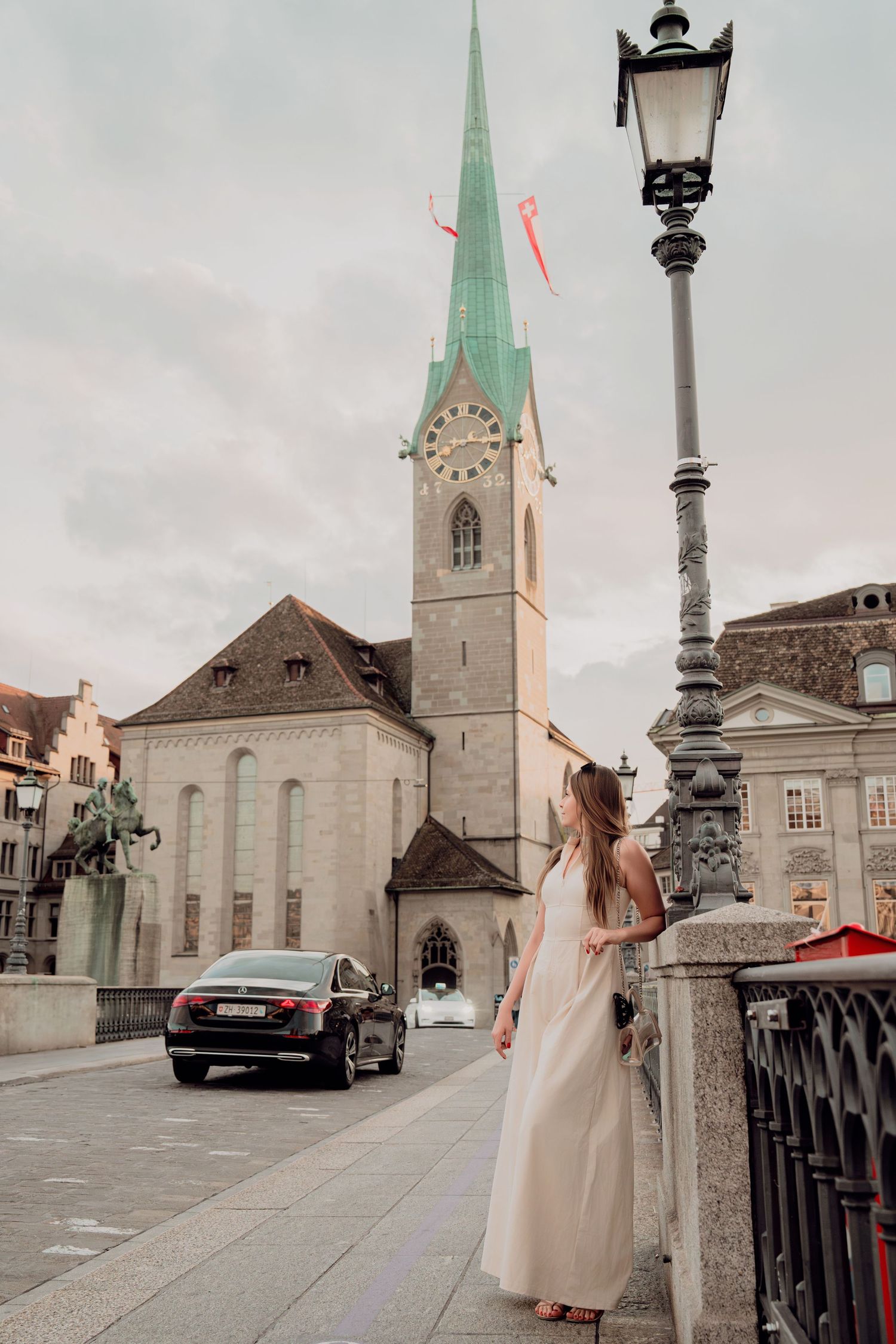 Woman in white dress stands on Munsterbrucke bridge near vintage lamppost with Fraumunster Church, Zurich.