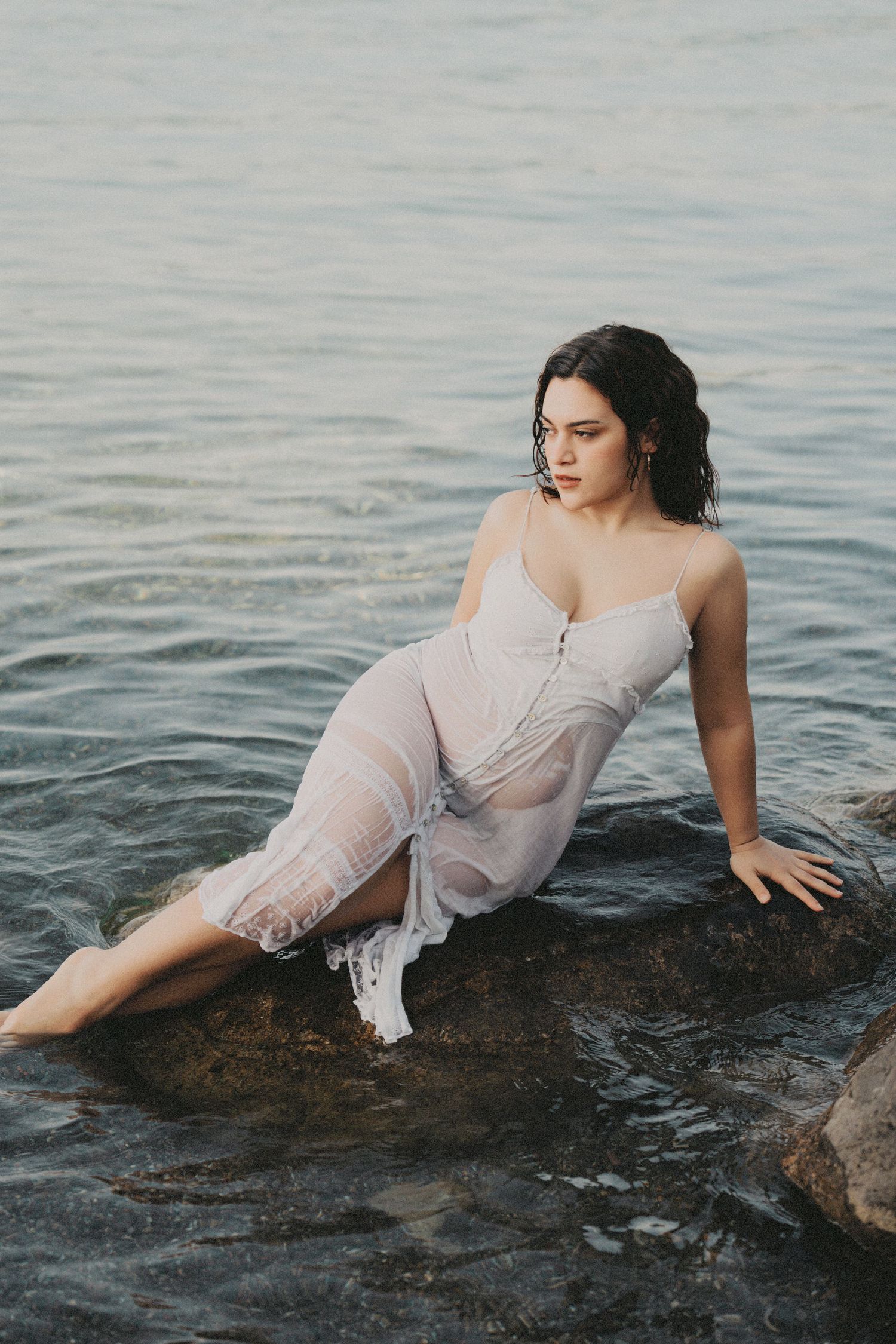 Woman in wet white dress sitting on submerged rocks in shallow water.